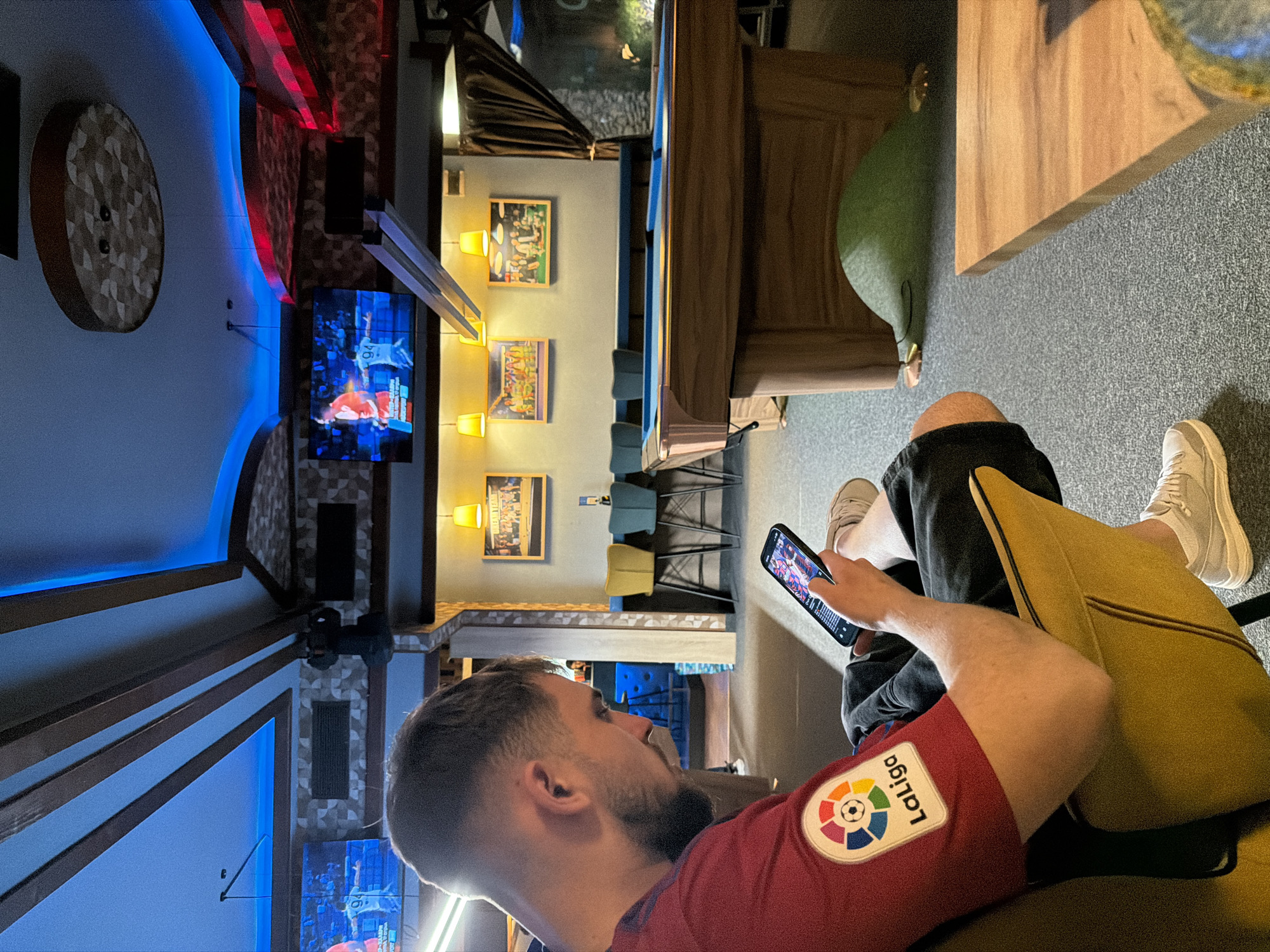 A man sits indoors, looking at his phone in Plovdiv, Bulgaria. He is wearing a red shirt with a "LaLiga" patch. Behind him are wall decorations and a TV showing a sports game. There is a pool table nearby. He has on two different shoes, one looks off-white and the other looks white.