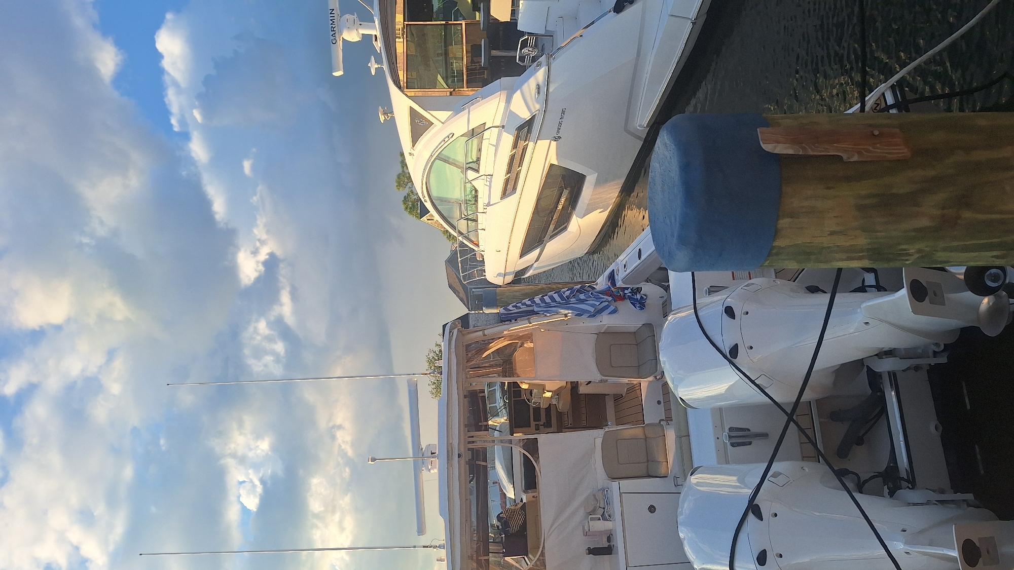 The image captures a close-up view of two boats docked in a harbor, likely in Nantucket, Massachusetts, USA, during the late afternoon or early evening. The sky above is filled with dramatic, cumulus clouds, some illuminated by the warm, setting sun, casting a golden glow on the scene.
In the foreground, the stern of a white boat is visible, equipped with twin white outboard motors. The boat appears to be moored to a wooden pilothad a blue protective cover. A black rope runs diagonally across the outboard motors.
To the right of this boat is a sleek, white motor yacht with a distinctive, curved windshield and dark tinted windows. The yacht is also docked and appears to be well-maintained. A radar dome labeled "GARMIN" is visible on the roof of a structure attached to the yacht.
Between the two boats, a blue and white striped towel is draped over a wooden post. In the background, several houses with light-colored siding and dark roofs are visible, suggesting a waterfront residential area or a marina. The water is dark and calm, reflecting the sky and the boats.
There are no people visible in the image. The overall impression is one of a peaceful, serene evening at a harbor.