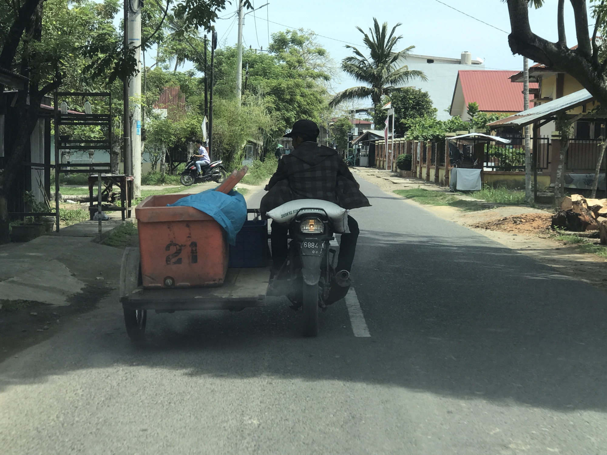 A rear view captures a person riding a motorcycle with an attached sidecar or utility trailer along a paved road. The driver is wearing a dark plaid jacket and a cap. A white bag, partially obscuring the license plate, is strapped to the motorcycle seat. The license plate is visible as "6884 M" with "HONDA" faintly above it. The trailer, made of metal, carries a large orange container with the number "21" stenciled on its side, and some blue fabric or plastic material.
The road appears to be a two-lane asphalt surface, with a faint white line visible near the center. Lush green trees and palm trees line both sides of the road, indicating a tropical or subtropical setting. Utility poles are present along the left side. In the background, another person on a motorcycle is visible further down the road. Several houses and simple structures with fences and red-tiled roofs are scattered along the roadside. The overall scene suggests a daytime setting, likely sunny, given the bright illumination. The environment appears to be a rural or semi-urban area in Lhokseumawe, Indonesia.