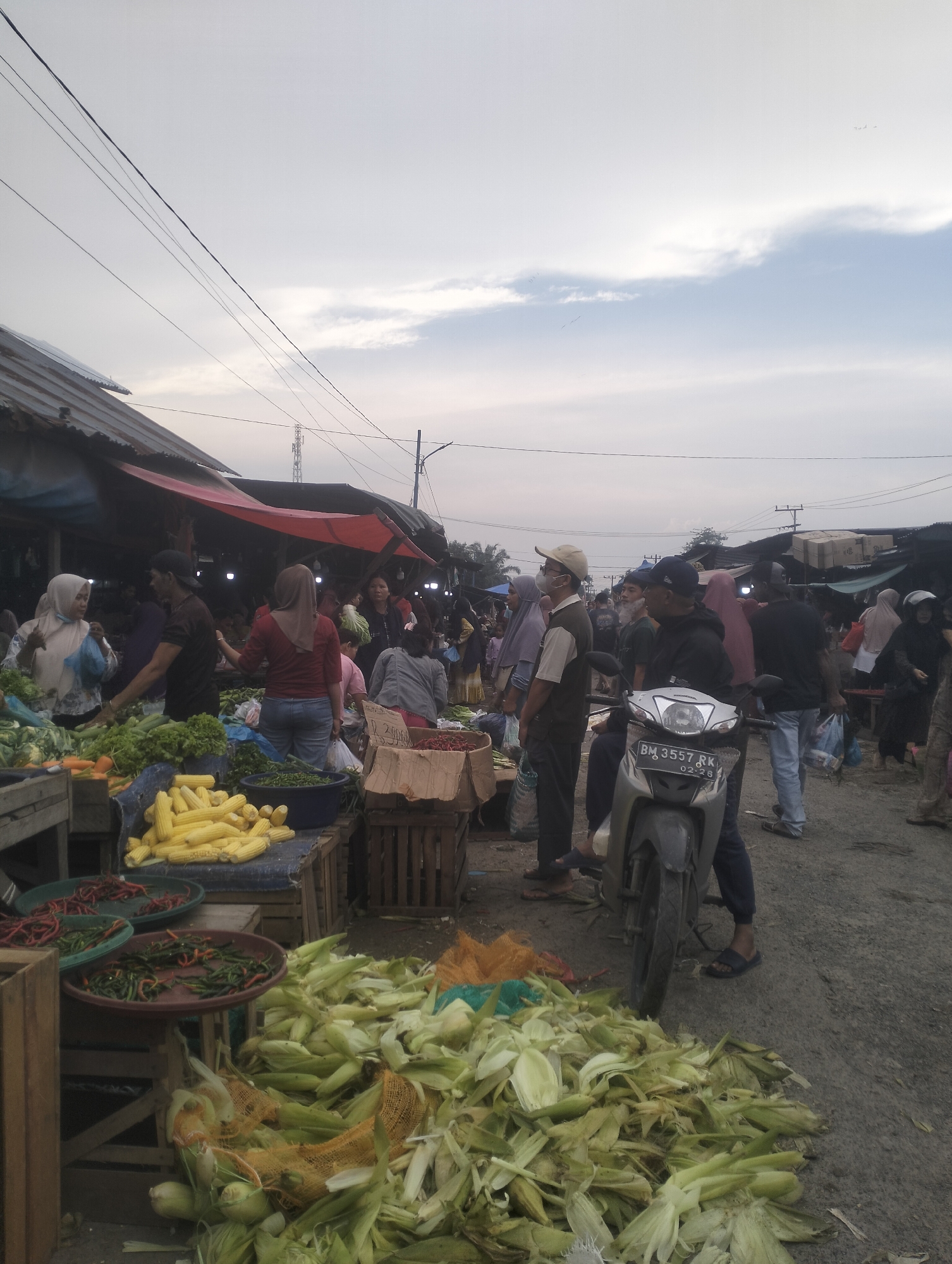 This image depicts an outdoor market scene in Dumai City, Indonesia, likely during the late afternoon or early evening given the lighting conditions. The market appears to be a produce market, with stalls selling various vegetables and fruits. In the foreground, a large pile of corn husks and discarded corn cobs are visible on the ground, suggesting recent sales or preparation. To the left, several crates and baskets contain fresh produce, including corn on the cob, chili peppers, and leafy green vegetables.
Numerous people are present in the market, engaged in various activities. Some are browsing the produce, while others appear to be vendors attending to their stalls. A motorcycle is parked prominently in the middle ground, suggesting a mode of transport used by either a customer or vendor. The background shows more market stalls with red awnings, and the sky is overcast with some clouds. Utility wires are strung across the upper portion of the image, a common sight in urban and semi-urban Indonesian settings. The overall atmosphere is one of a bustling local marketplace.