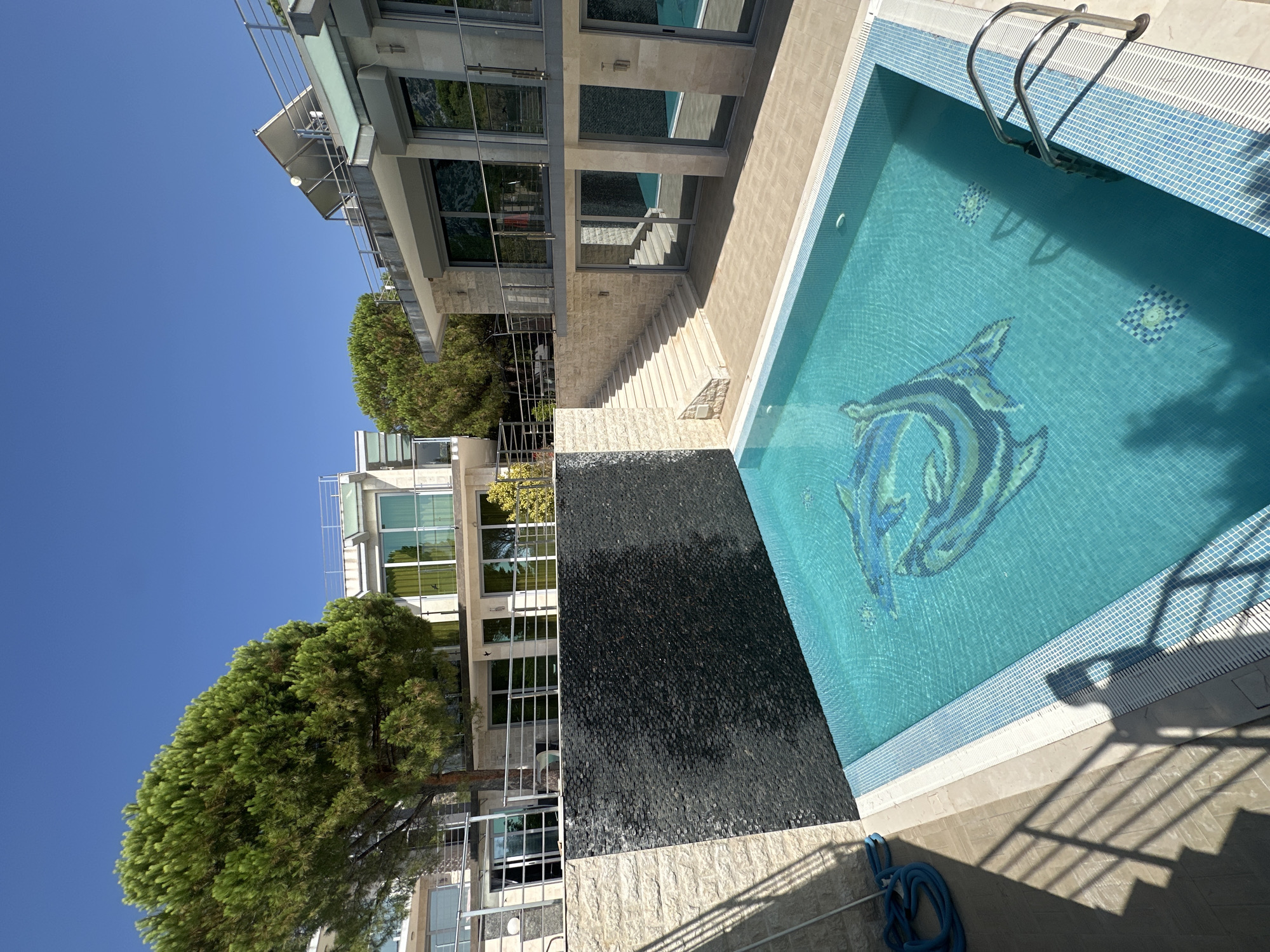 The image shows a bright, sunny day in Brca, Montenegro, with a focus on a modern residential property featuring a swimming pool. In the foreground, a rectangular swimming pool with turquoise water is visible. The pool bottom is decorated with a mosaic of two blue and green dolphins. The pool's edge is lined with white and blue tiles, and a silver ladder provides access to the water. To the left of the pool, a dark, textured wall, possibly a waterfall feature, is set against a light stone wall. A blue hose is coiled on the ground near the pool.
Behind the pool, a paved patio area leads up to a multi-story building with a contemporary design. The building has large glass windows, balconies with metal railings, and light-colored stone facades. One section of the building features an external staircase made of light stone. A large, mature pine tree with dense green foliage stands to the left of the building, providing shade. In the background, more of the modern buildings are visible, along with lush green trees and a clear blue sky. The overall scene suggests a luxurious and well-maintained residential estate in a coastal or hilly area.