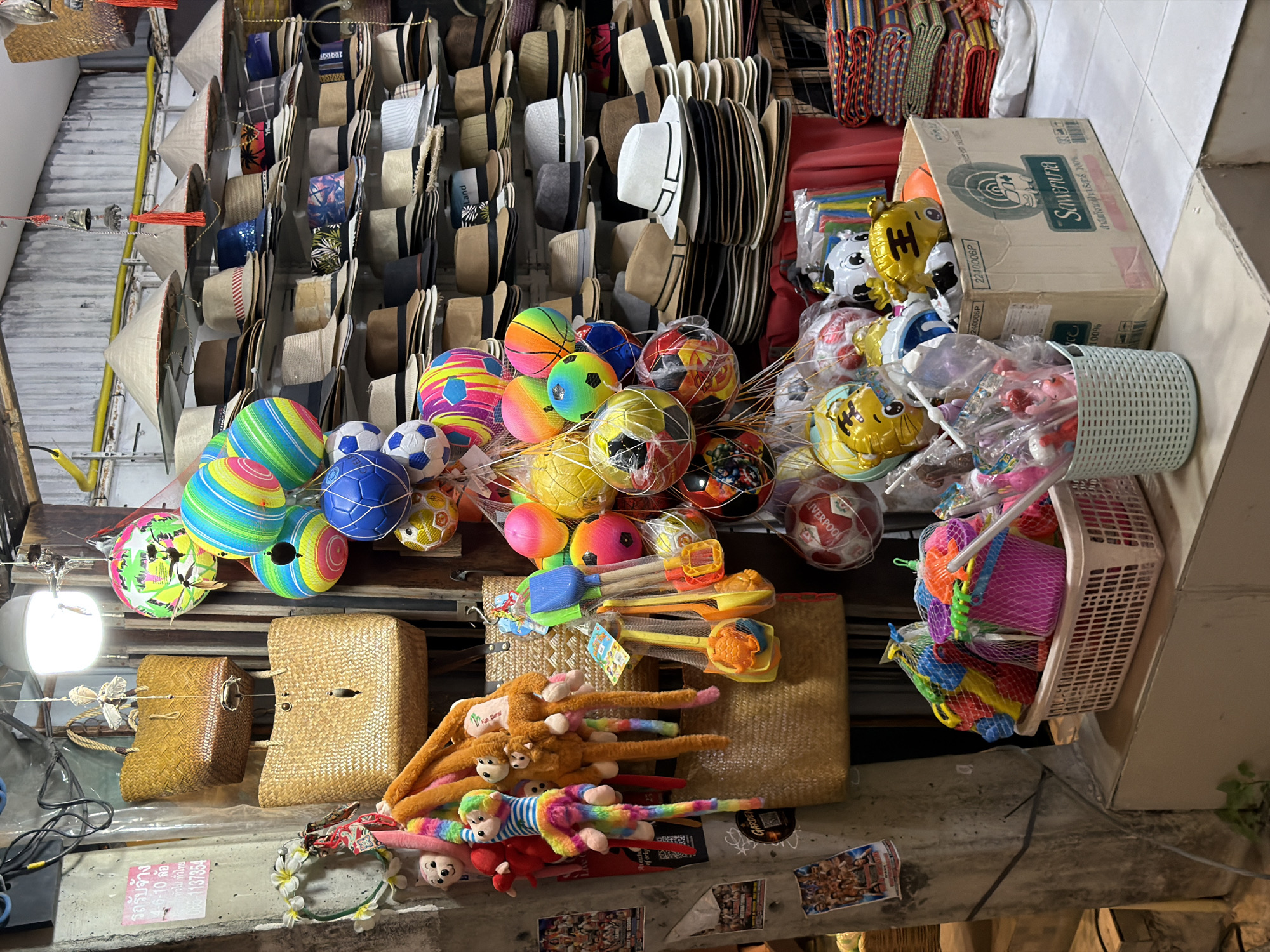 The image depicts a market stall in Baan Bo Phut, Thailand, overflowing with a variety of goods. In the foreground, a white plastic basket holds a collection of brightly colored children's beach toys, including shovels and buckets. To the right, a light blue woven basket contains more toys and packaged candy. Behind these, a cardboard box is filled with inflatable balloons and various plastic toys. A stack of colorful woven mats is visible on the far right.
Dominating the middle ground is a tiered display of hats, featuring a mix of fedoras, straw hats, and conical Asian hats, arranged neatly on shelves. Hanging from above are numerous colorful, spherical objects resembling decorative balls or large ornaments. Also hanging are several plush toys, including monkeys and bears, attached to a woven bag. Two large woven straw bags are prominent, one hanging above the other. Various posters and flyers are affixed to a rough concrete pillar on the left side of the stall. The overall impression is one of a vibrant and crowded retail environment characteristic of a local market.