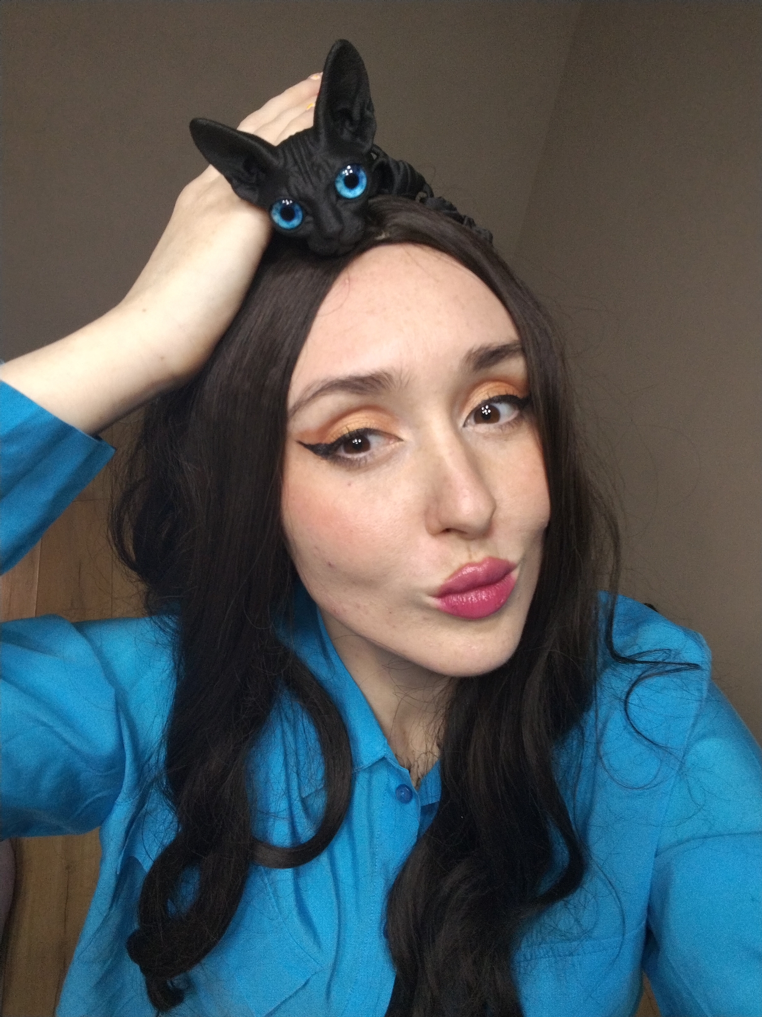 A woman poses for a selfie, wearing a blue collared shirt and a full face of makeup including winged eyeliner and pink lipstick. She is holding up her left hand towards her head. A black cat with blue eyes is perched on top of her head. The background appears to be a neutral-toned wall. The image provides no evidence that it was taken in Izhevsk, Russia.