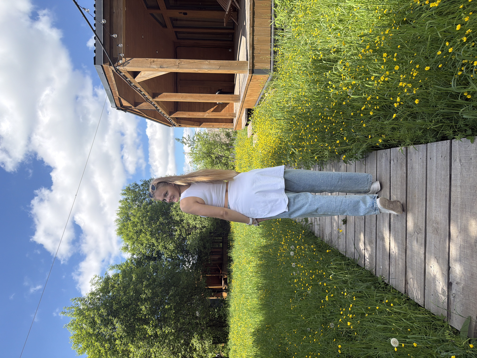 The image, taken in Vozhoiskii, Russia, captures a bright outdoor scene on a sunny day. A young woman with long blonde hair, wearing a white sleeveless crop top, light blue jeans, and light-colored sneakers, is the main subject. She stands on a wooden walkway, looking slightly to her left with a relaxed expression and a subtle smile, her sunglasses resting on her head.
The walkway stretches from the foreground into the distance, running alongside a vibrant field of lush green grass densely covered with small yellow flowers, resembling buttercups. Sunlight illuminates the scene, casting subtle shadows on the wooden planks of the path and the surrounding foliage.
In the background, a rustic wooden gazebo or open-sided structure with a dark roof stands amidst the greenery. Decorative string lights are visible along the eaves of the gazebo. Beyond this structure and the field of flowers, a line of dense green trees frames the horizon, beneath a clear blue sky dotted with fluffy white clouds. The overall impression is one of a peaceful and pleasant outdoor setting, likely a park or recreational area, during the daytime.