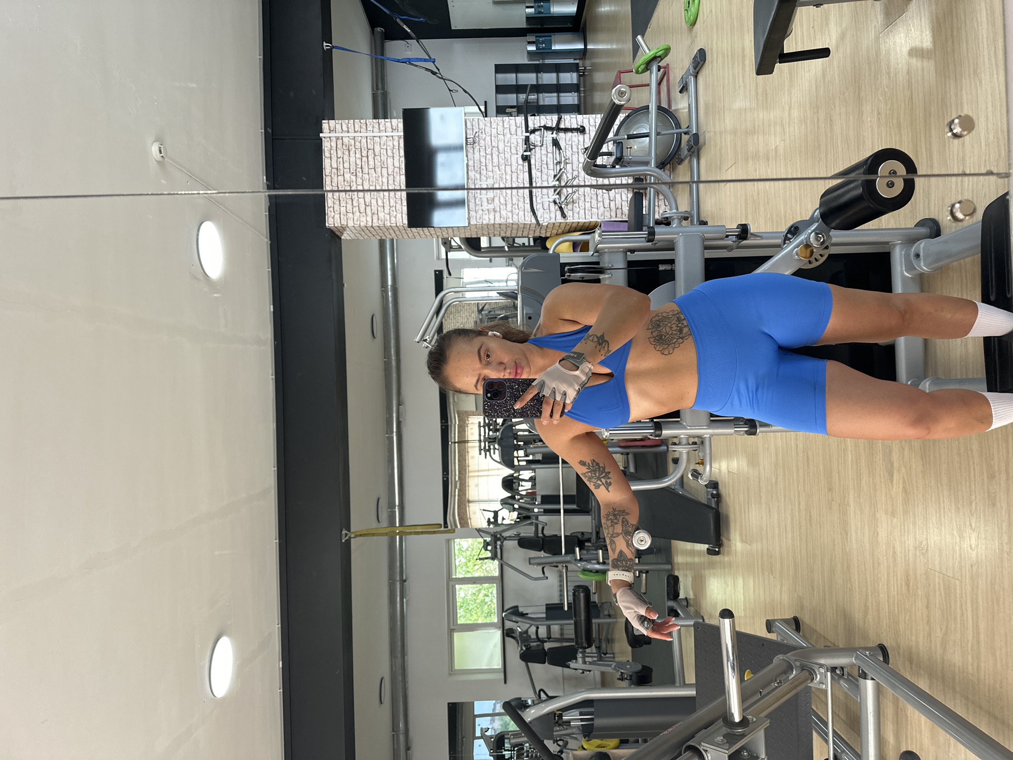 The image presents a forensic view of a female subject taking a mirror selfie within a gym setting. The gym interior features light wooden laminate flooring, white walls with a dark grey horizontal band near the ceiling, and a light-colored ceiling with recessed circular light fixtures and a white smoke detector. Visible on the ceiling are faint, elongated streaking patterns, possibly indicative of water damage or repairs. The scene, according to the prompt, is located in the city of Yuzhnouralsk, Russia.
The subject is a woman, appearing in her mid-20s to early 30s, dressed in athletic attire consisting of a royal blue sports bra and matching high-waisted cycling shorts. She wears light grey fingerless workout gloves and white athletic socks. A white wireless earbud is visible in her left ear, and a white smartwatch is on her left wrist. Her dark hair is pulled back. The woman displays several tattoos: a large floral design on her upper right arm, two distinct floral tattoos on her left forearm, and a large, intricate floral or mandala-like tattoo on her lower back, partially visible above the waistband of her shorts. She holds a black smartphone with a textured black case in her right hand, angled towards the mirror to capture her reflection. Her expression is neutral.
The background, seen through the mirror's reflection, reveals a variety of gym equipment, including grey-framed strength machines with black padding, a black workout bench, and a machine with a green weight plate. A prominent rectangular pillar with faux-brick textured wallpaper stands in the mid-ground, featuring a black flat-screen television mounted on its side. In the far background, large windows show green foliage outside, indicating a daytime setting. Other gym elements include a water cooler with a blue dispenser, black shelving units, and yellow and blue suspension training straps hanging from the ceiling. A vertical seam or edge of the mirror is visible, running through the center-right of the frame.