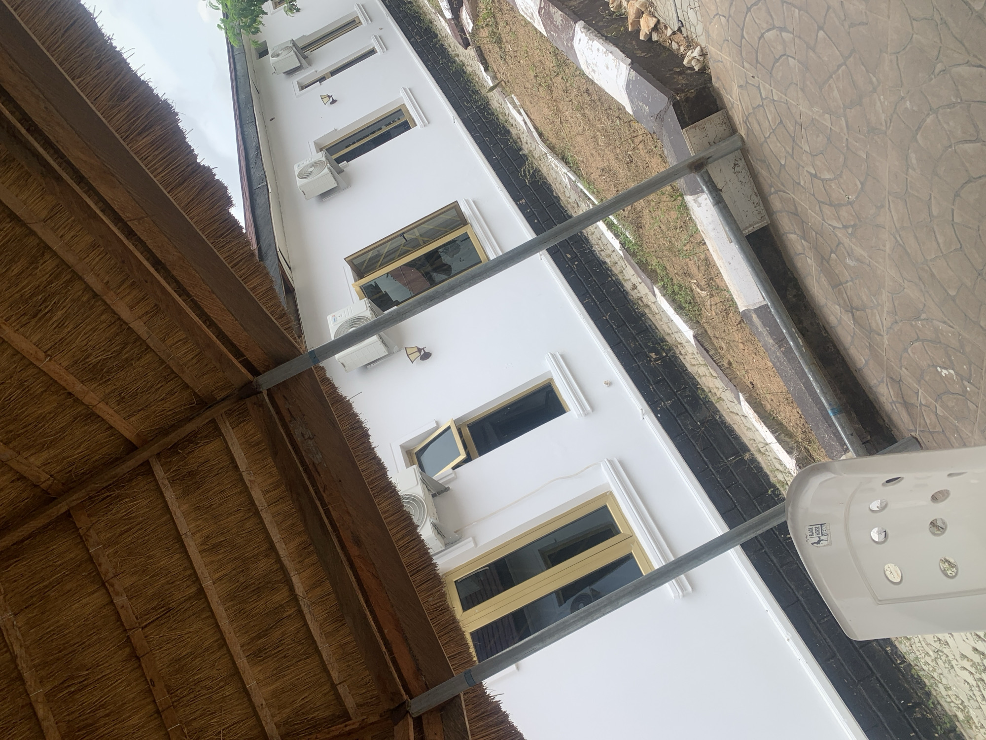 The image captures a close-up view of a white building with multiple windows and air conditioning units, located under a thatched roof structure. The building's exterior features a black and white striped foundation, and the windows have golden frames. In the foreground, a white plastic chair is visible, with a sticker on its back that reads "BLACK HORSE EVENT". The ground area appears to be a mix of dirt and paved tiles. The scene is set in Jalingo, Nigeria.