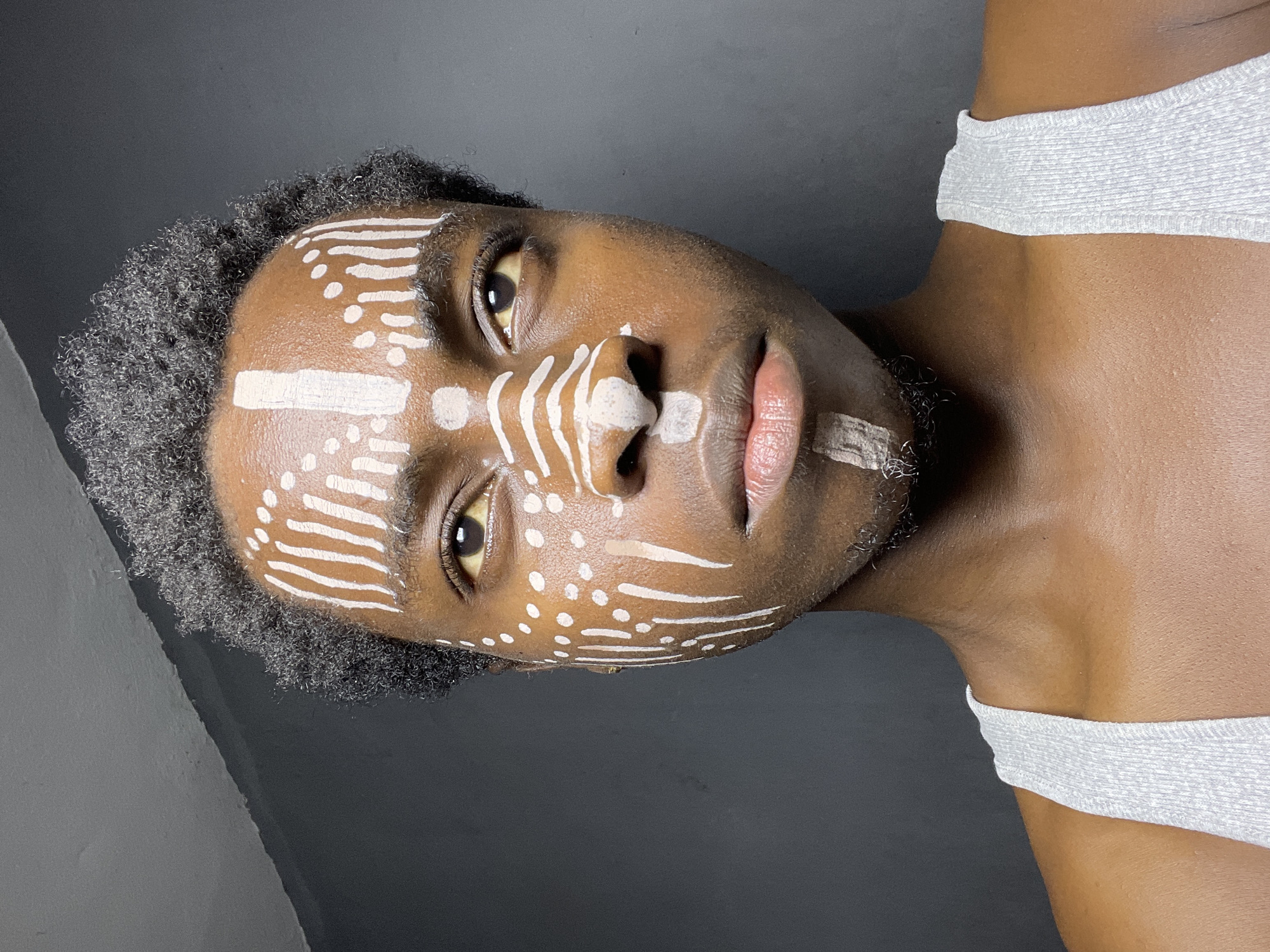 A close-up portrait shows a dark-skinned person with a neutral expression and white paint designs on their face. The designs include lines and dots across their forehead, cheeks, and nose. Their hair is short and dark. They are wearing a light gray top. The background is a plain, dark gray color. The image is slightly tilted. The location is not discernible from the image.