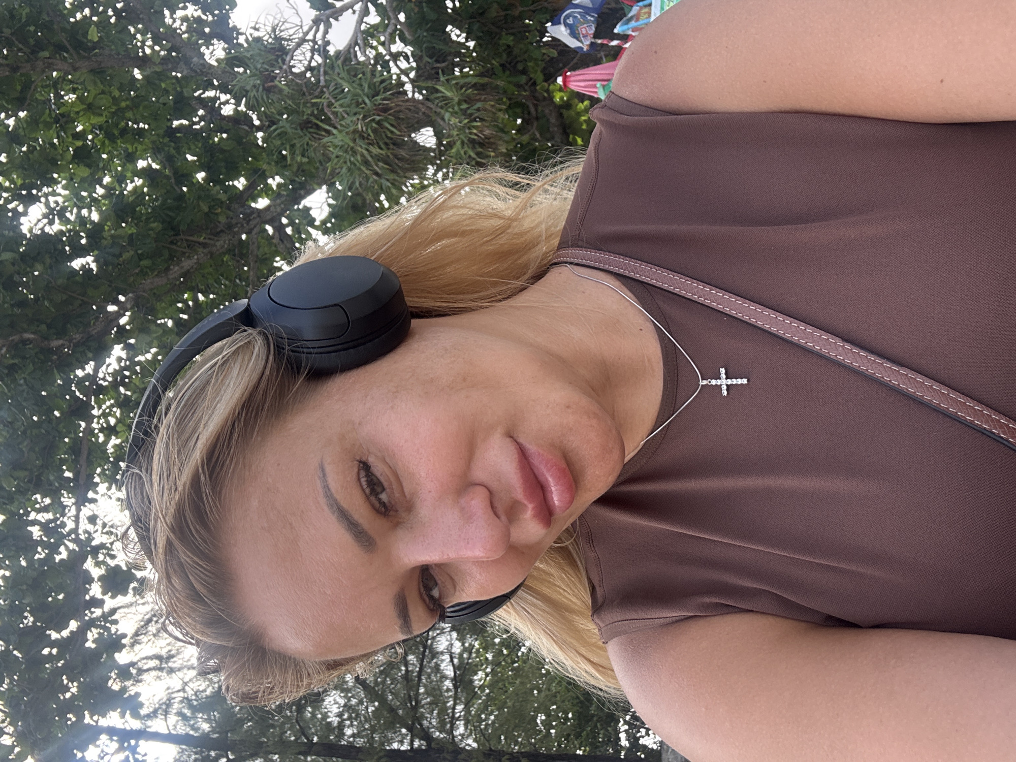 A woman with blonde hair and headphones is taking a selfie outdoors. She is wearing a brown top and a silver cross necklace. In the background, there are trees and beach umbrellas. The location is the city of Patong, Thailand.