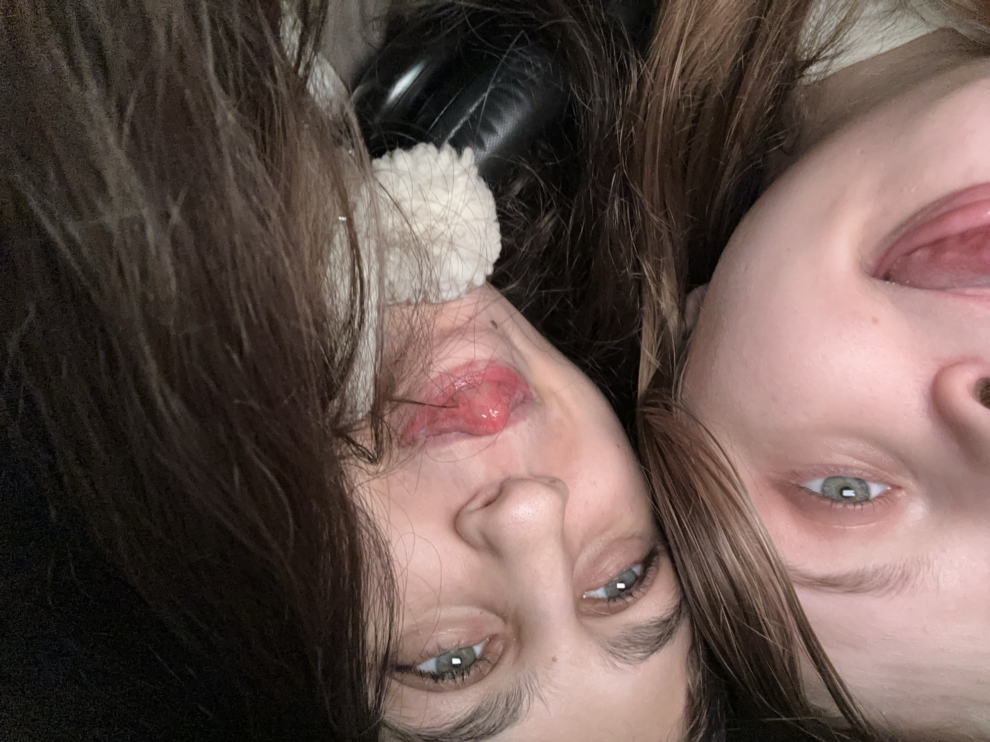 This close-up, rotated image captures two young individuals, likely females, in a playful pose. Their faces are positioned intimately close together, filling the frame. Both have fair skin and light blue-grey eyes. The person on the left (when the image is rotated for upright viewing) has dark brown hair and a slight smile, with her tongue partially visible. A white, fluffy, pom-pom-like object, possibly an accessory or part of clothing, is visible near her ear and chin. The person on the right has medium brown hair and is actively sticking her tongue out in a clear expression of playfulness. Both individuals' eyes are directed towards the camera. The background is indistinct, composed primarily of their hair and dark, soft materials, suggesting an indoor setting. The activity is a casual, lighthearted interaction, likely a selfie or a close-up snapshot. No specific location cues are visible within the image itself to confirm Moscow, Russia, nor is any text present. The time of day is indeterminate due to the indoor lighting.
