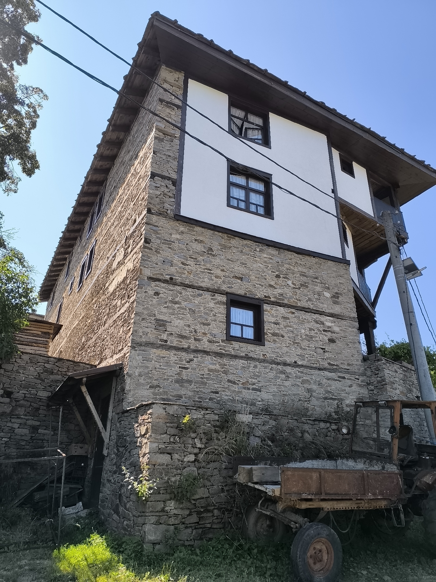 An eye-level, wide shot captures a traditional stone and plaster house with a wooden-tiled roof in the Bulgarian city of Kovachevitsa. The house, with its rough-hewn stone lower levels and whitewashed upper stories, features multiple dark-framed windows. Overhead, power lines stretch across a clear blue sky, and to the left, the branches of a leafy tree reach into the frame. In the foreground, a rusty, abandoned tractor sits partially obscured by overgrown grass and weeds. The scene is devoid of people.