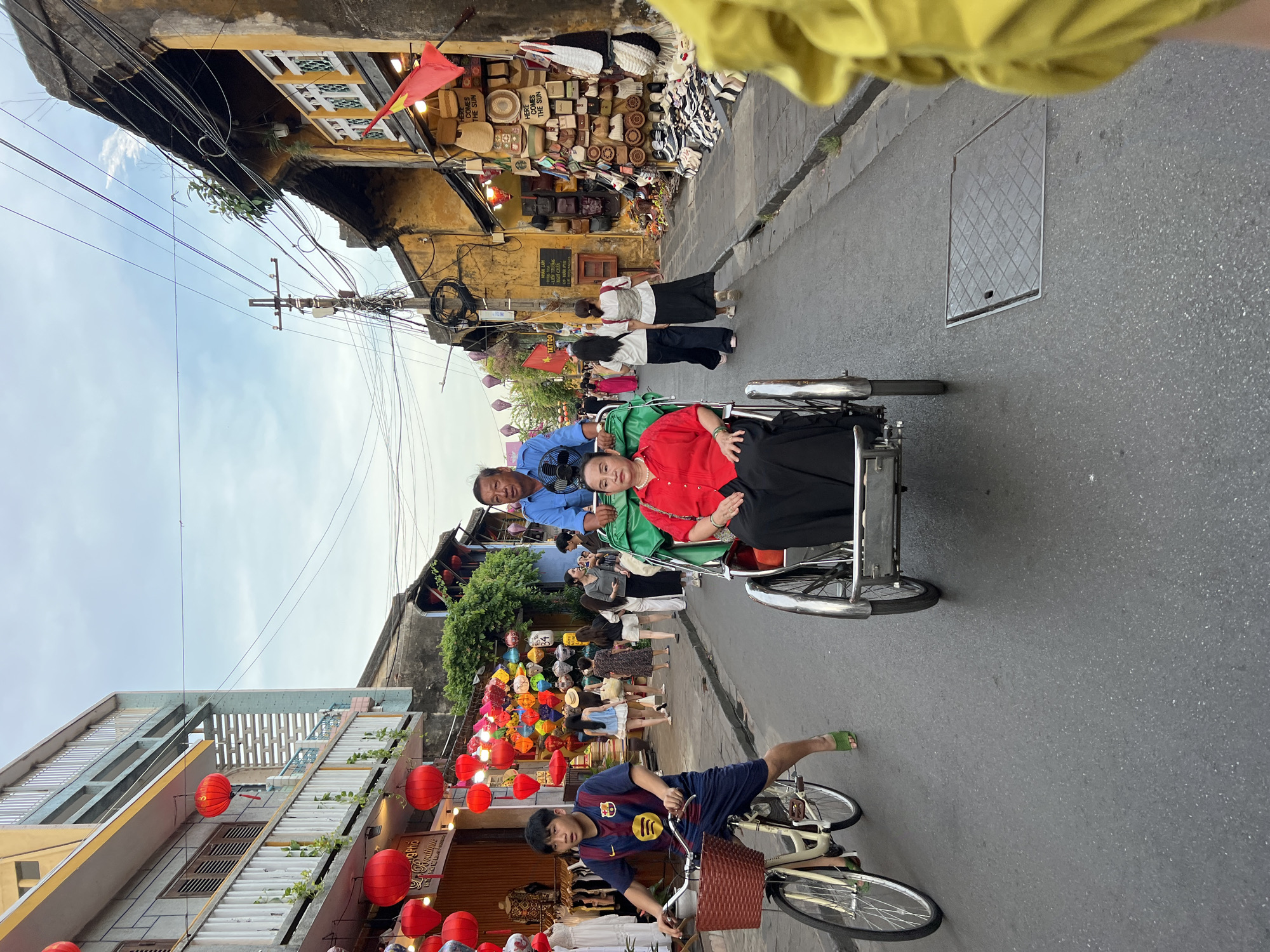 The image shows a street scene in Hội An Ward, Vietnam. In the foreground, a young person rides a bicycle. Behind the cyclist, a cyclo carries two passengers, an elderly woman and a man, who is operating the cyclo. The street is lined with buildings, many of which are adorned with red lanterns. The buildings exhibit a traditional architectural style, with yellow facades and wooden shutters. Shops selling various goods, including bags and clothing, are visible along the street. The sky is overcast, and the overall atmosphere suggests a bustling street in a tourist area.