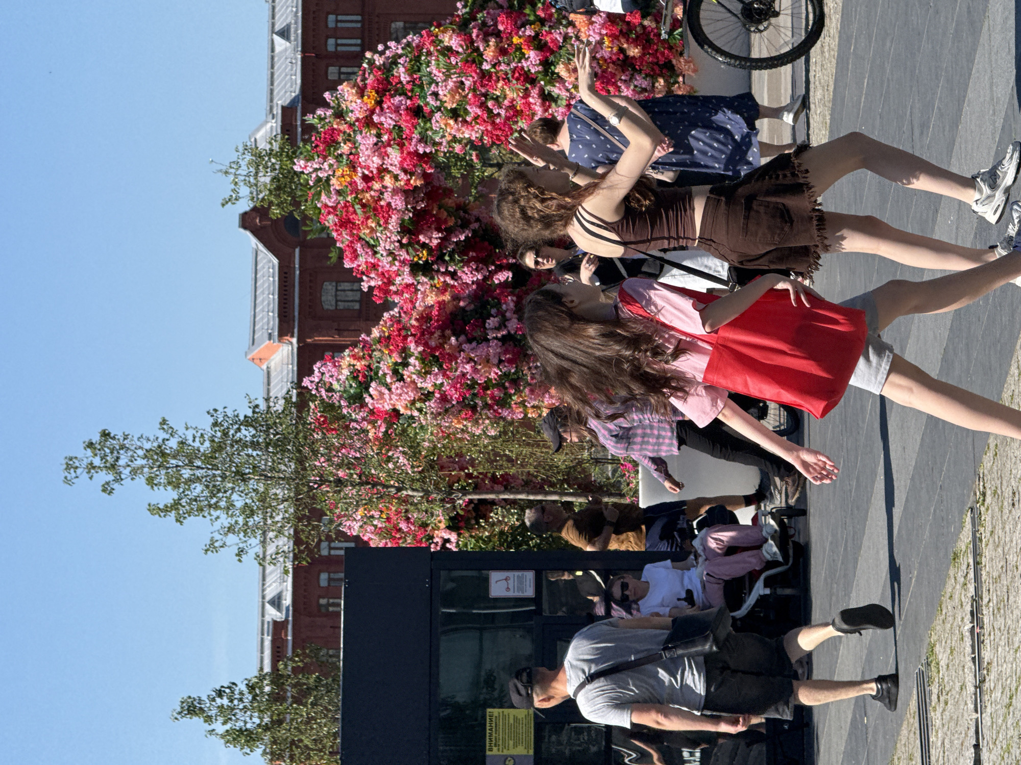 The image depicts a bustling outdoor scene on a sunny day in Zaryadye Park, Moscow, Russia. The sky is clear and blue, indicating fair weather. In the background, a large red-brick building with a grey metallic roof and multiple windows is visible, partially obscured by green deciduous trees. Dominating the midground are two immense, arch-like floral installations, densely covered in vibrant pink, red, and orange blossoms, creating a festive atmosphere.
In the immediate foreground, two young women are walking from left to right across a paved pathway. The woman on the left has long brown hair and is wearing a light pink short-sleeve top, grey shorts, and carrying a red tote bag on her left shoulder. The woman on the right has curly brown hair and is dressed in a brown cropped tank top with thin straps, a brown mini-skirt with a frayed hem, and white athletic shoes. Her arms are raised and outstretched, suggesting she is actively speaking or gesturing.
To the left of these women, a dark-colored kiosk or structure is present, featuring reflective panels and a visible yellow sign with the Cyrillic word "ВНИМАНИЕ!" (ATTENTION!) and a scooter icon, confirming the Russian language and likely indicating rules regarding scooter use.
Several other individuals populate the scene. Further left, a man is walking away from the viewer, wearing a grey t-shirt, dark shorts, a black baseball cap, and a black messenger bag. Next to the kiosk, a woman in a white t-shirt and pink pants is seated in a black wheelchair or stroller. To the right of the kiosk, a man in a brown shirt and dark shorts walks left, followed by a woman in a plaid shirt and black baseball cap. Further right, partially obscured by the foreground figures and the floral arrangements, a woman in a blue polka-dot dress walks. A partial view of a bicycle wheel is visible on the far right. The ground consists of grey pavers with visible metal grates. The overall scene conveys a lively public space filled with people enjoying the sunny weather and the striking floral decorations.