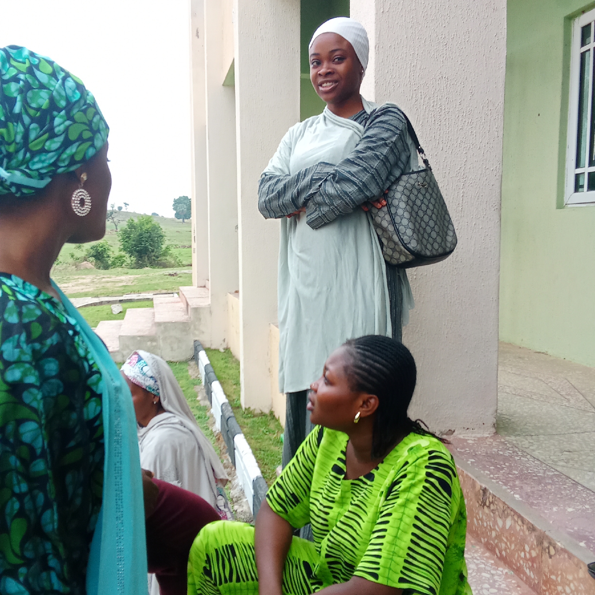 Four women are present in an outdoor location in Akwanga, Nigeria. One woman, standing near a beige column, is fair-skinned, wearing a white head covering and light-colored garment, and has a patterned bag over her shoulder. She is smiling. Another woman, partially visible in the foreground, wears a green and blue patterned headwrap and matching garment, with large circular earrings. A third woman, seated on steps, wears a bright green garment with black stripes and small hoop earrings. The fourth woman is partially visible, seated, and wearing a light-colored headscarf and light-colored top.
Behind the women are steps, a black and white guardrail, and a green hillside. A building with a green-painted section and a window is visible to the right.