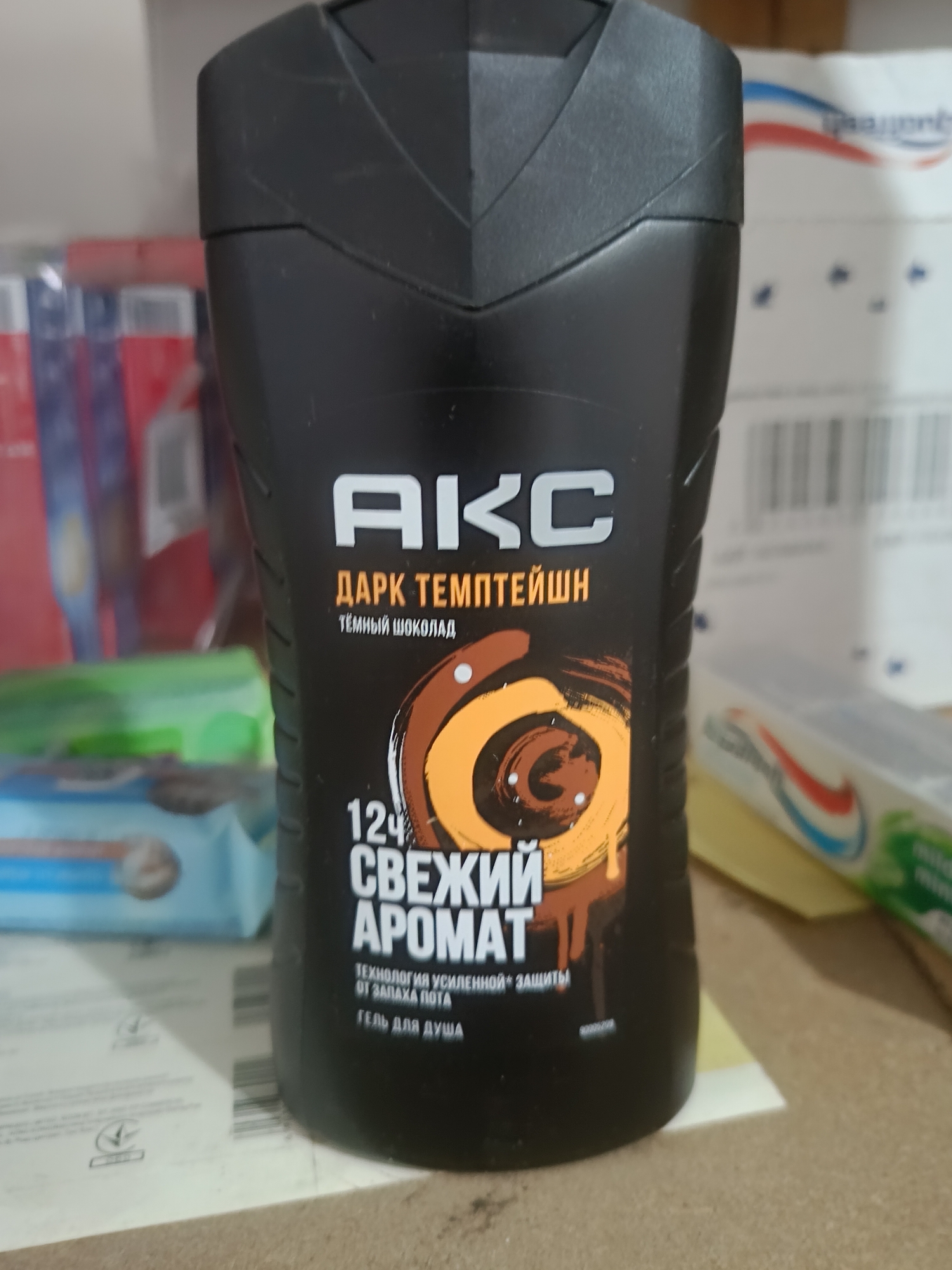 A black bottle of Axe Dark Temptation shower gel is the primary subject of the image. The bottle has a textured cap and features prominent white lettering spelling out "AKC" followed by "ДАРК ТЕМПТЕЙШН" (Dark Temptation in Russian) in orange text. Below this, smaller text reads "тёмный шоколад" (dark chocolate). A graphic of a swirling chocolate design is visible on the front of the bottle. The text "12ч СВЕЖИЙ АРОМАТ" (12h Fresh Aroma) is also clearly displayed in white. Smaller text below indicates "ТЕХНОЛОГИЯ УСИЛЕННОЙ* ЗАЩИТЫ ОТ ЗАПАХА ПОТА" (Enhanced* Protection Technology Against Sweat Odor) and "ГЕЛЬ ДЛЯ ДУША" (Shower Gel). The bottle is positioned on a flat surface, possibly a table or shelf. In the background, out-of-focus elements suggest other products and packaging, including what appears to be a box with a red, white, and blue stripe design, and possibly some disposable razors. The lighting is somewhat dim, casting shadows. The overall scene is consistent with a product shot or a retail environment. There is no information in the image to suggest a specific location in the city of Berdiansk, Ukraine.