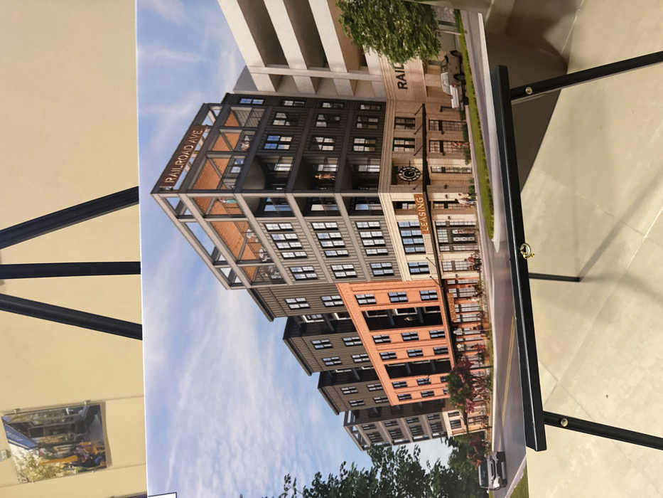 An architectural rendering displays a modern building complex. "RAILROAD AVE" is prominently displayed on the building. A clock and a "LEASING" sign are visible on the facade. The structure features a mix of materials including brick, metal, and glass. The rendering includes sky and trees, suggesting a completed development within its surroundings. The artwork is displayed on an easel.