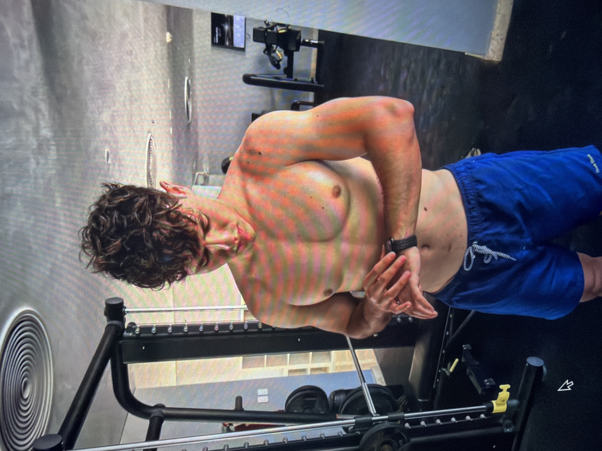 The image displays a young adult male, shirtless, within an indoor gym setting, purportedly located in Shumen, Bulgaria. The man stands with his body slightly angled to the left, his head bowed, focusing on a dark smartwatch worn on his left wrist. Both of his hands are positioned over the watch, suggesting interaction or adjustment. He possesses a muscular physique with visible chest definition, broad shoulders, and developed biceps. His hair is dark, curly, and styled upwards. He wears plain blue athletic shorts with a white drawstring.
The gym environment features professional-grade equipment. To the left, a large black multi-station cable machine or Smith machine is prominent, showcasing vertical guide rods with numbered adjustment holes and a yellow locking pin near the base. Behind this, several black weight plates are stacked. The floor is dark, consistent with gym flooring. In the background, other exercise machines are partially visible, including what appears to be a dark cardio machine on the right. The walls are light-colored, and the ceiling features multiple circular air diffusers or vents, along with recessed lighting fixtures. A dark monitor or television screen is mounted on a wall in the mid-background to the right.