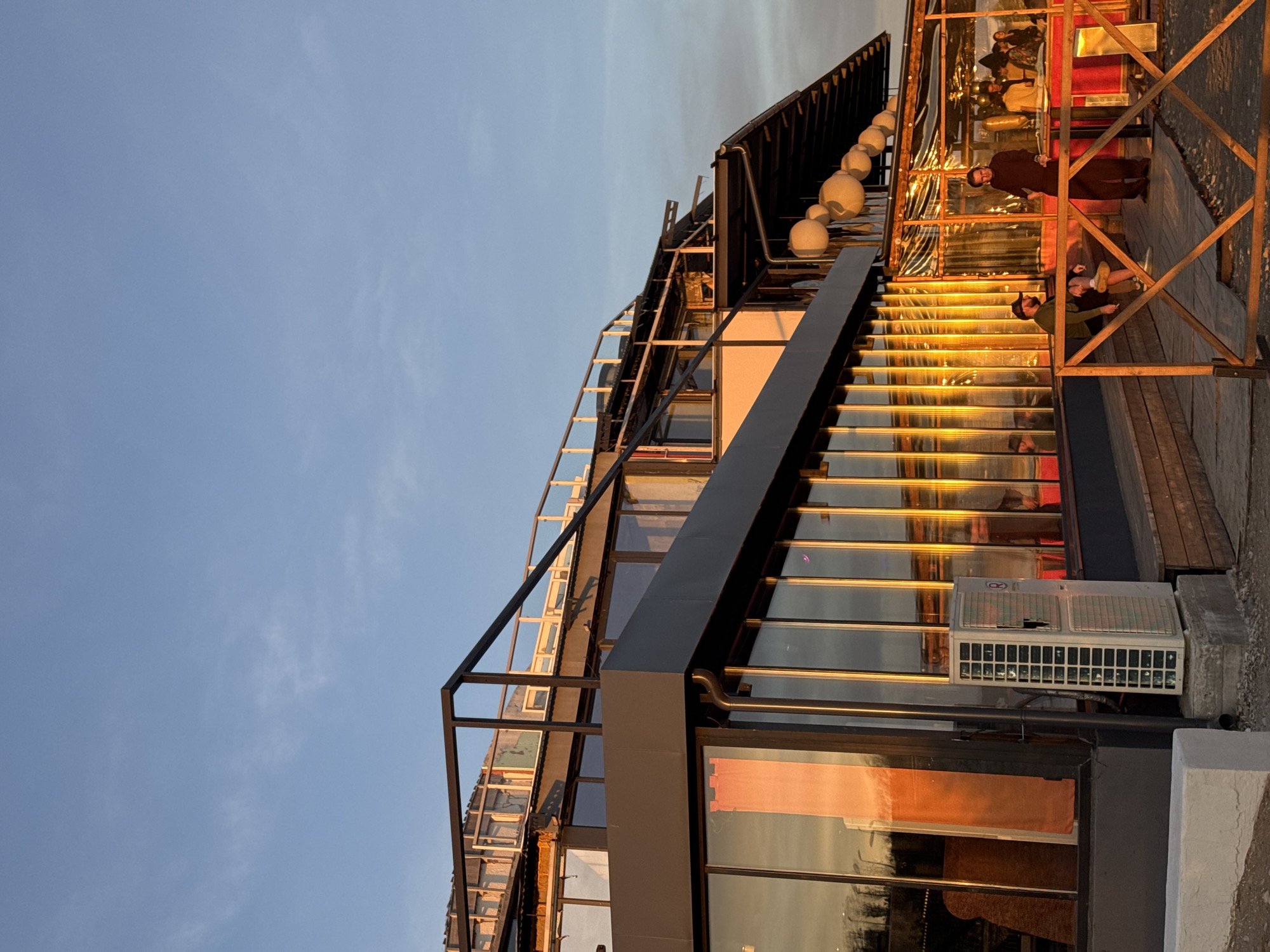 The image shows a modern, multi-level building in an urban environment under a clear blue sky. The building has a geometric design with numerous windows and glass panels, reflecting the warm sunlight. A prominent staircase with illuminated steps ascends along the building's facade. The architectural style appears contemporary, with a combination of metal framework and glass surfaces.
On the top level, there are a few people present. Two individuals appear to be seated or lounging, while one person is standing and facing in their direction. The lighting suggests that it is either early morning or late afternoon, contributing to the ambiance.