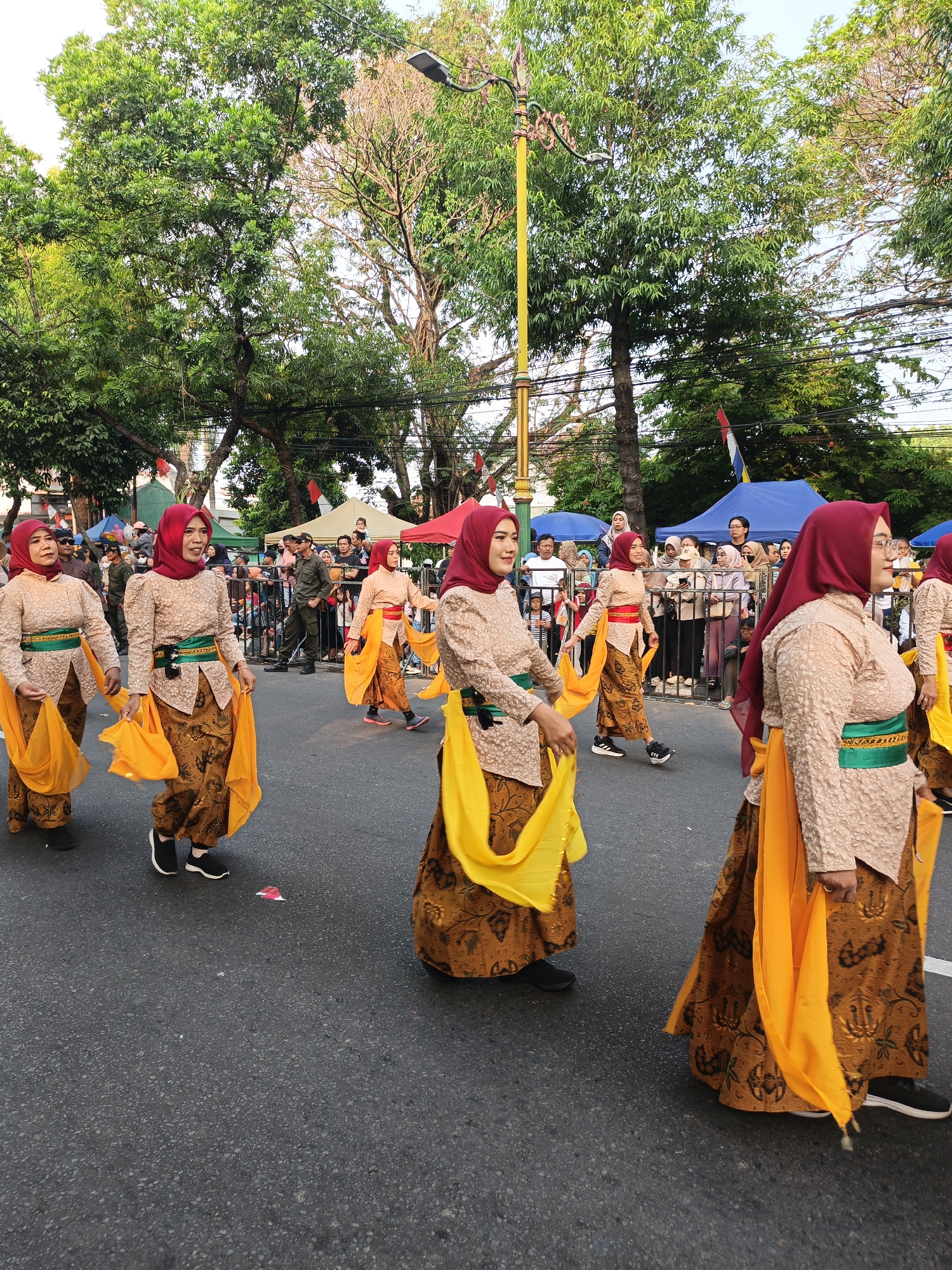 The image depicts a street parade in Gayamprit, Indonesia, featuring a group of women in traditional Indonesian attire, specifically a kebaya and batik skirt. They are wearing red hijabs and holding vibrant yellow sashes. The women are walking in a line, seemingly performing a dance or a processional. In the background, there are spectators, tents, and trees lining the street, indicating an outdoor event. The atmosphere appears festive and cultural.