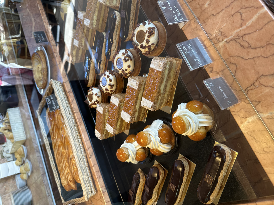 The image depicts an interior view of a patisserie display case, specifically at a Café Carette establishment in Paris, France. The perspective is slightly elevated, looking down through a clear glass partition at an array of French pastries.
In the immediate foreground, a dark, reflective surface holds numerous individual pastries. On the left, three chocolate éclairs, each with a small label indicating "LE CAFE CARETTE," rest on golden rectangular bases. To their right, two St. Honoré cakes are visible, characterized by a circular base, whipped cream, and small caramel-glazed choux buns; one is marked with a "St Honoré" price tag for 8€. Adjacent to these are five rectangular slices of mille-feuille, displaying multiple layers of pastry and cream, with one carrying a "Mille-Feuille" price tag for 7.50€. Throughout this section, several round pastries, topped with white frosting and dark chocolate pieces or nuts, are present on dark bases. One such pastry is near a tag indicating "Tarte à la noix de Pécan" for 8.50€. Additional chocolate éclairs extend into the mid-ground.
Above this primary display, a reddish-brown marble counter supports further items. A large, braided golden pastry, possibly an apple turnover, is presented in a woven wicker basket and priced at 4.80€. To its right, a round fruit tart rests on a white plate. In the upper left background, stacked white ceramic plates and smaller pastries are visible, out of sharp focus. Reflections on the display glass suggest an indoor, brightly lit environment. No individuals are clearly visible, though blurred reflections hint at activity beyond the frame.