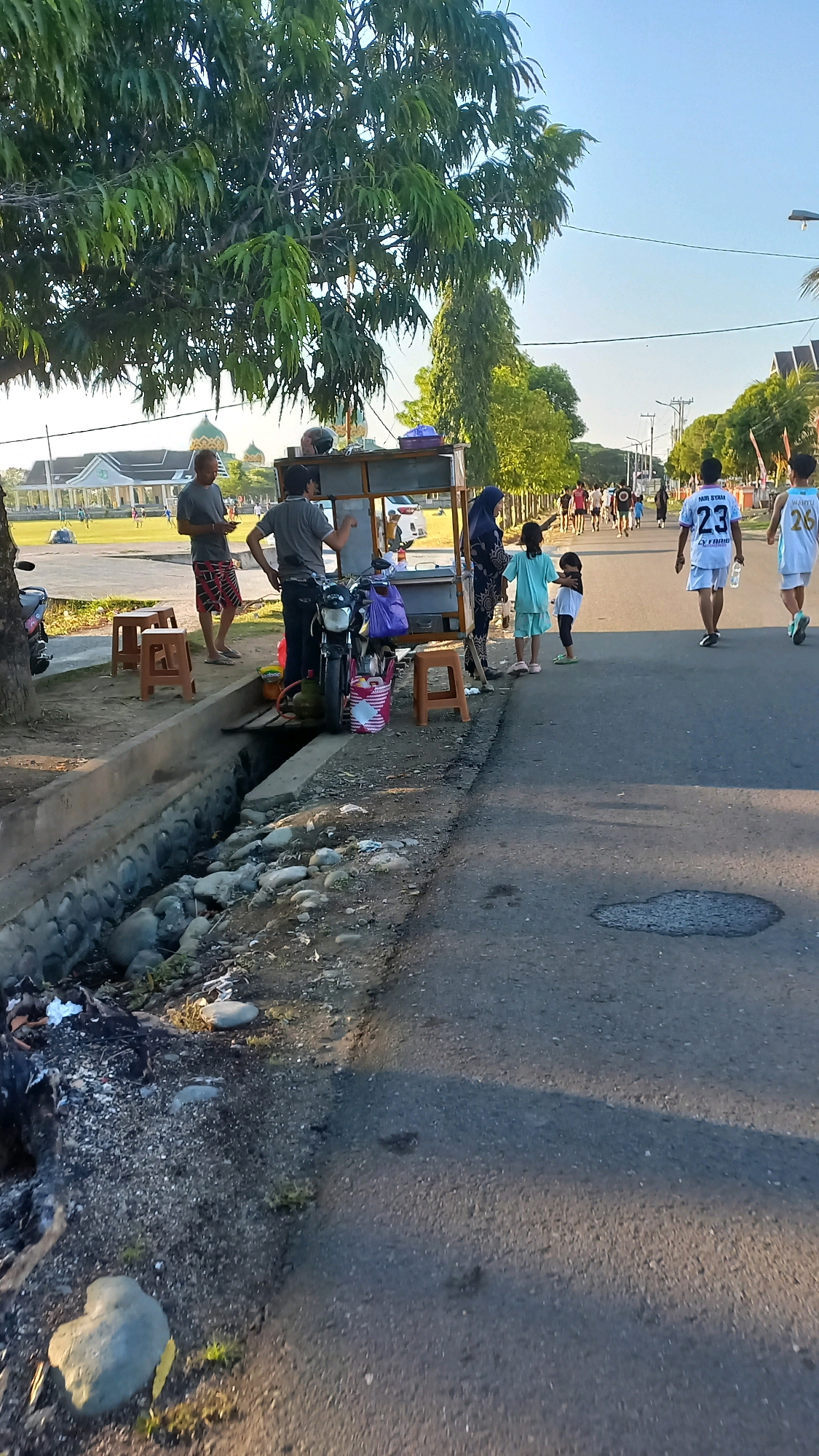 In the city of Sabe, Indonesia, a street scene unfolds under a bright, clear sky, likely in the early morning or late afternoon given the long shadows. To the left, a group of people are gathered around a food stall, indicated by a cart with a canopy and various containers. A motorcycle is parked near the stall. In the background, a building with domed roofs and a sports field are visible, suggesting proximity to a community or recreational area.
Further down the street, a group of individuals, predominantly young males, are walking or running on the paved road. Some are wearing athletic attire, with numbers visible on their shirts, such as "23" and "26". A woman and two young children are also present near the food stall, interacting with each other. The environment is lined with trees, and a concrete drain runs along the side of the road, containing water and debris. The road itself shows signs of wear, including a puddle or pothole. The overall atmosphere suggests a casual gathering or a local event taking place in a public space.