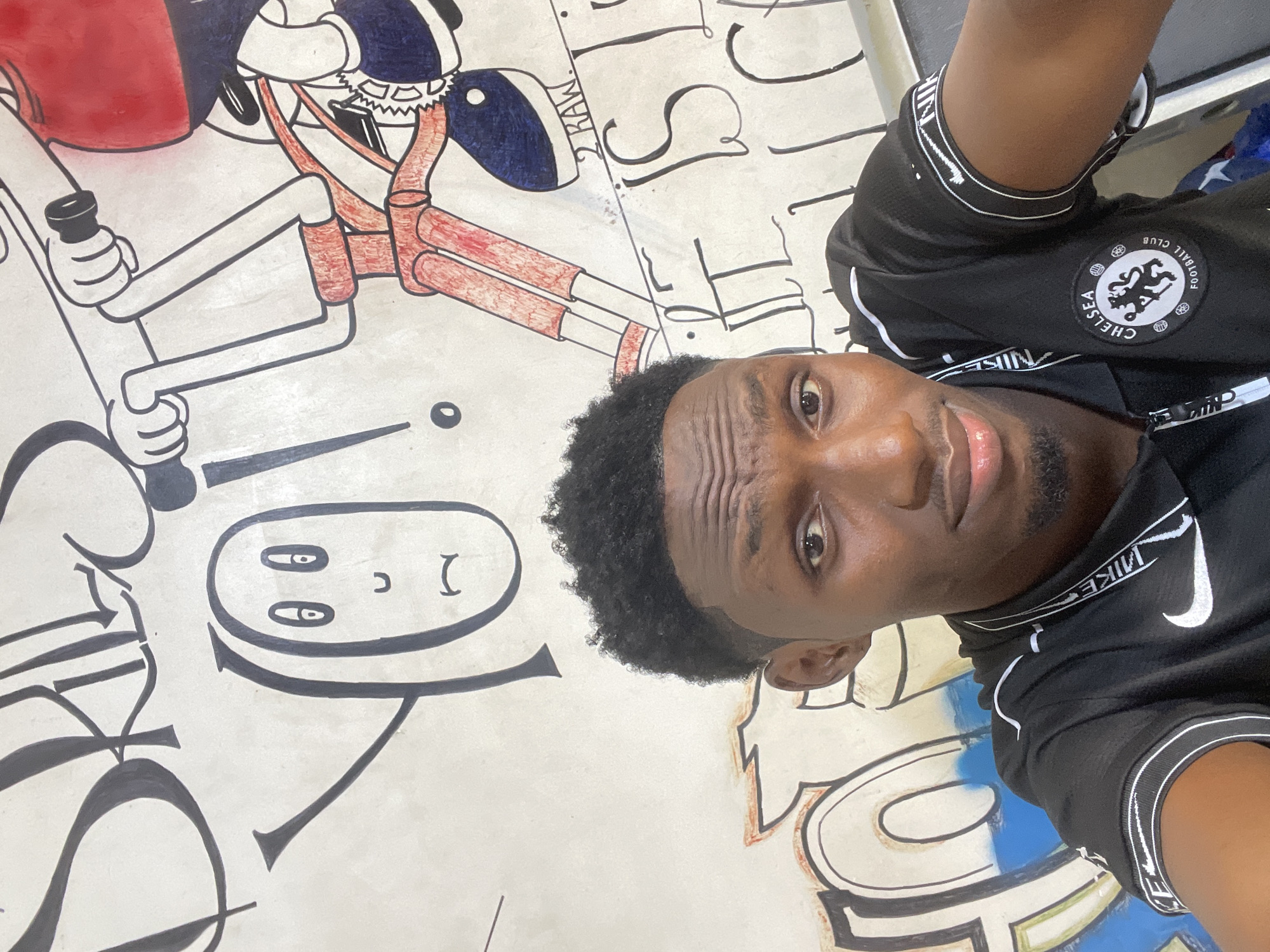A young Black male with dark skin, short, curly black hair, and a light beard is taking a selfie in front of a graffiti-covered wall. He is wearing a black polo shirt with white trim on the collar and sleeves, and a Chelsea Football Club logo is visible on the left side of his chest. The graffiti features stylized lettering, cartoonish figures, and bold lines in black, red, blue, and white. One prominent drawing depicts a character on a bicycle. The scene is likely indoors, given the lighting and the texture of the wall. This location is in Akowonjo, Nigeria.
