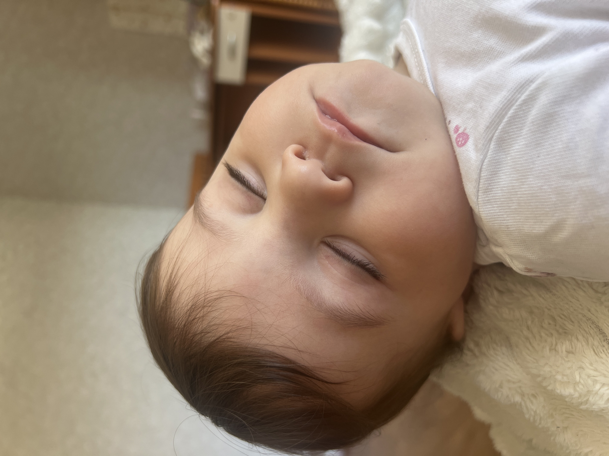 A close-up, angled portrait shows a sleeping baby with dark brown hair and closed eyes. The baby is wearing a white garment with a small pink strawberry design on the chest. The baby rests on a textured white surface, possibly a blanket. The background includes a blurred wooden structure, possibly furniture. There is no evidence to support that the location is in Elena, Bulgaria.
