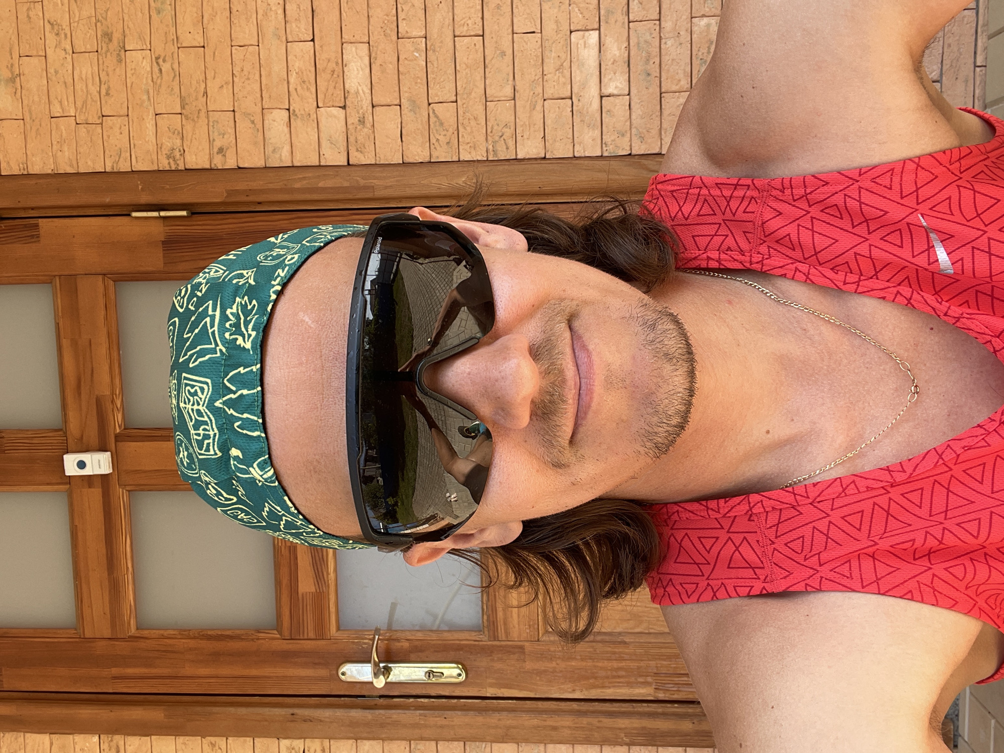 A man is wearing a green paisley headband, sunglasses, and a red Nike sports shirt. He is standing in front of a door with a brick wall above it. The door has a silver handle and lock. The man is wearing a gold necklace. The location is Перевозное, Russia.