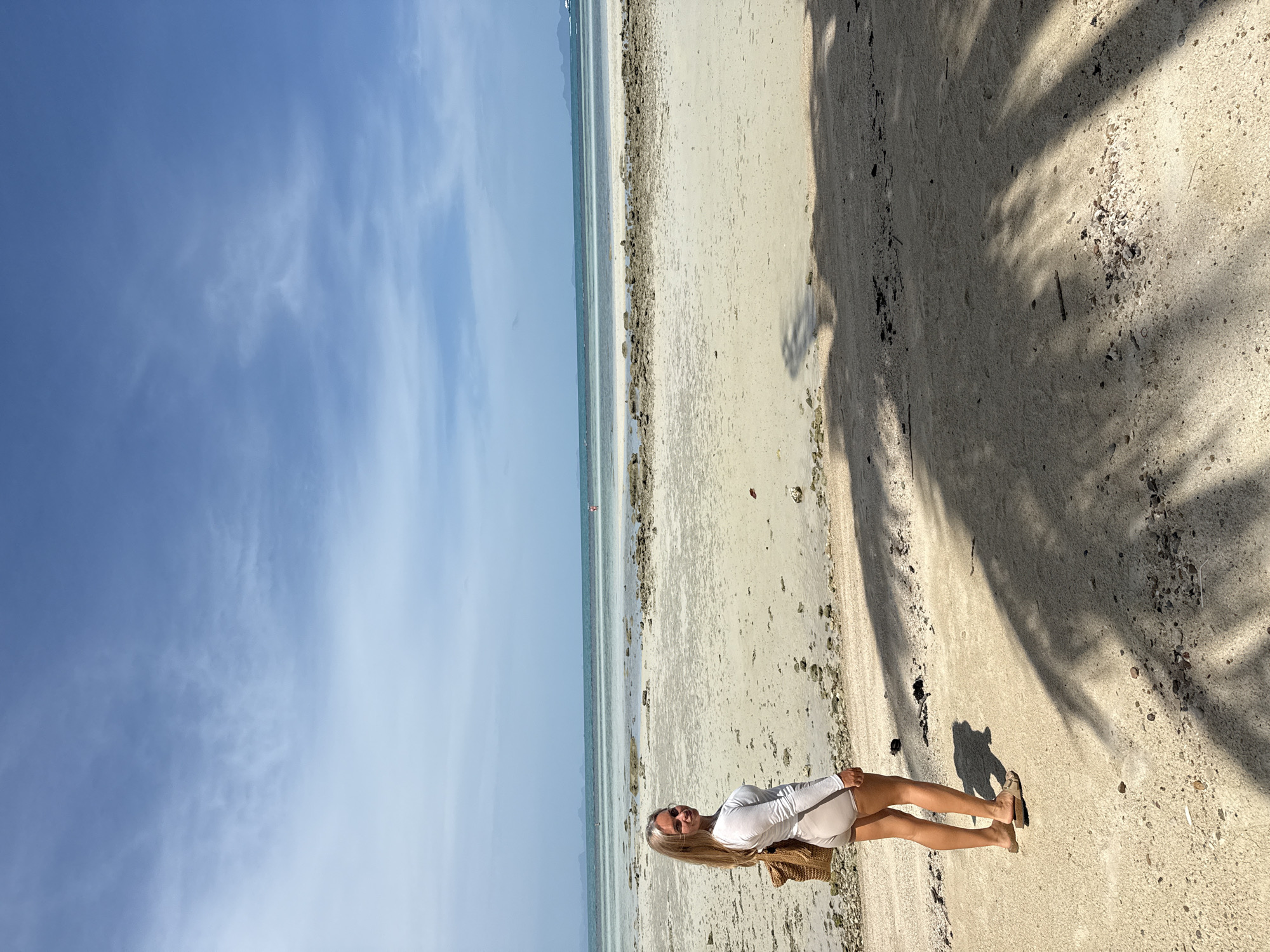 In Ko Samui District, Thailand, a woman stands on a sandy beach looking over her shoulder at the camera. She is wearing a white long-sleeved top and light-colored shorts, with a woven bag slung over her right shoulder and sandals on her feet. The beach is composed of sand and small pebbles, with scattered rocks and shells. In the distance, the ocean stretches out, with a faint horizon line where it meets the sky. The sky is a clear blue with sparse, wispy clouds. Palm tree shadows are cast across the sand in the foreground. The scene appears to be a bright, sunny day.
