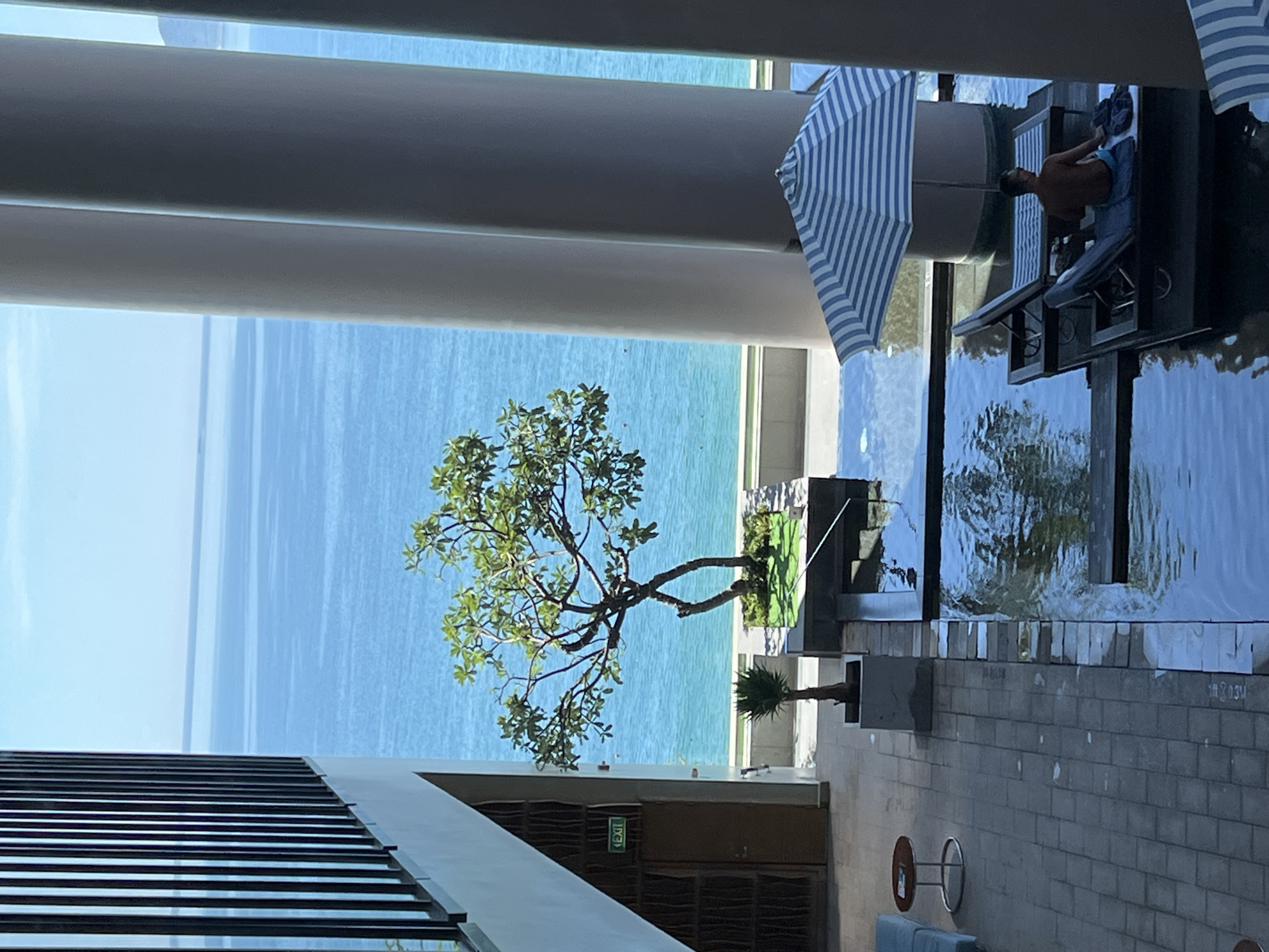 This is a medium-shot, eye-level photograph of a resort pool area overlooking the ocean in Nha Trang Ward, Vietnam. The scene captures a serene tropical environment with a man relaxing on a lounge chair in a shallow pool. The pool features a striped umbrella providing shade. The background is dominated by a vast expanse of blue ocean under a clear sky. To the left, a modern building with large columns and a balcony railing is visible, along with an "EXIT" sign above a door. A potted tree stands near the water's edge, adding a touch of greenery. The paving stones and the water's surface reflect the surrounding architecture and the sky, creating a sense of tranquility and luxury.