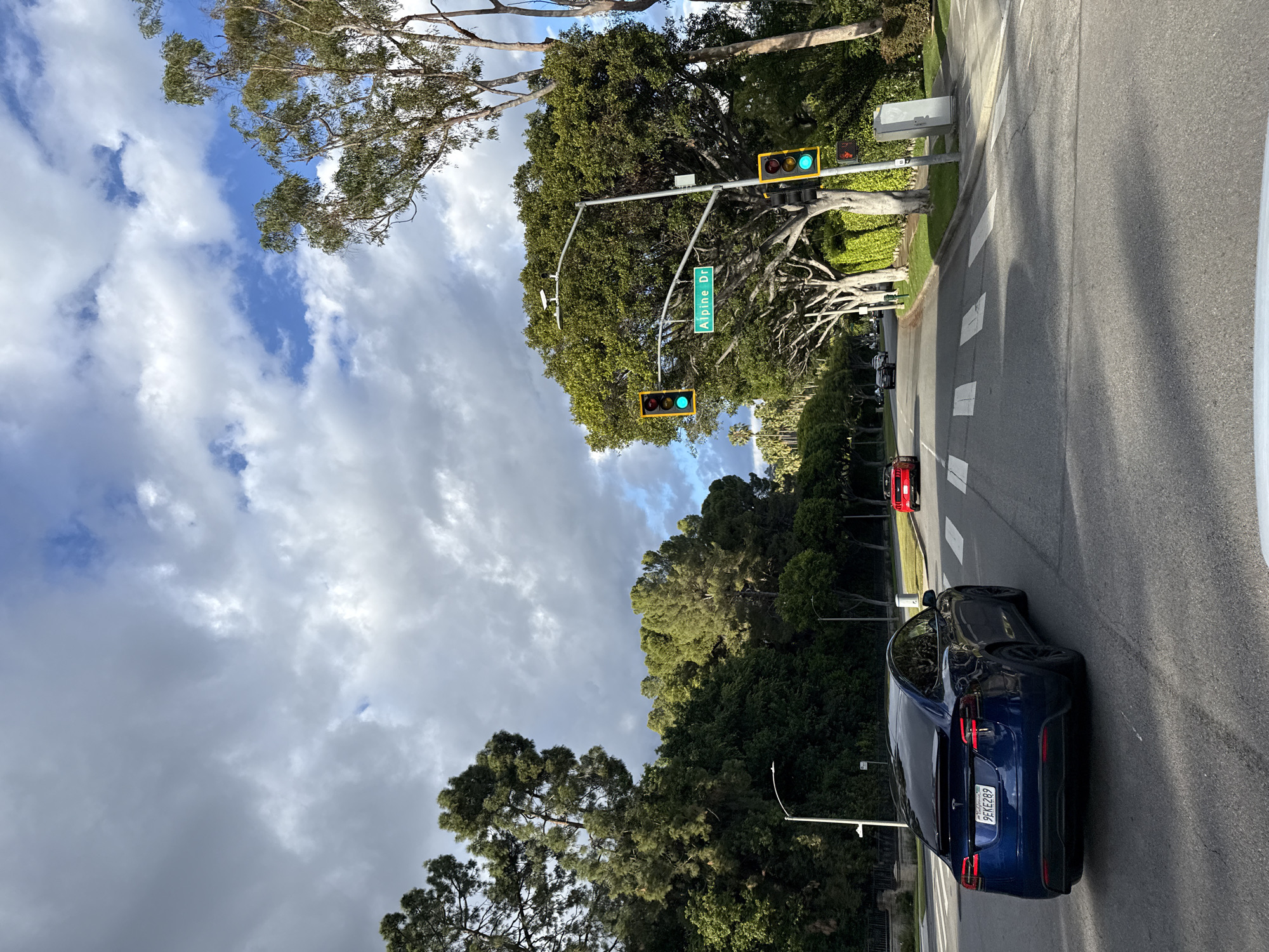 The image depicts a street scene in Beverly Hills, United States of America. The street is lined with trees and greenery. A dark blue Tesla SUV is parked on the right side of the road. The license plate reads "9EKE289". A red car is visible further down the street. The intersection shows a street sign indicating "Alpine Dr." The traffic lights are green. The sky is partly cloudy. There are pedestrian crossing lines at the intersection.