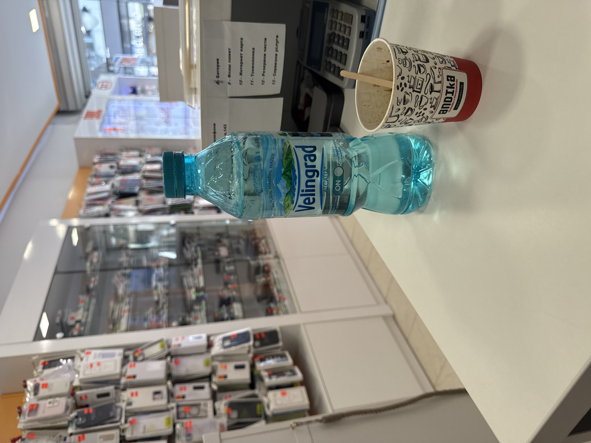 The image shows a tabletop scene with a bottle of "Velingrad" natural mineral water and a used paper cup with the "andika" logo on it. There's a wooden stick inside the cup. A calculator can be seen behind the bottle, and a list of services is taped to the nearby surface. In the background, there are display cases with phone cases, suggesting a store setting. The text on the list of services appears to be written in Bulgarian, suggesting the location is in Bulgaria. Burgas is not discernible in the image.