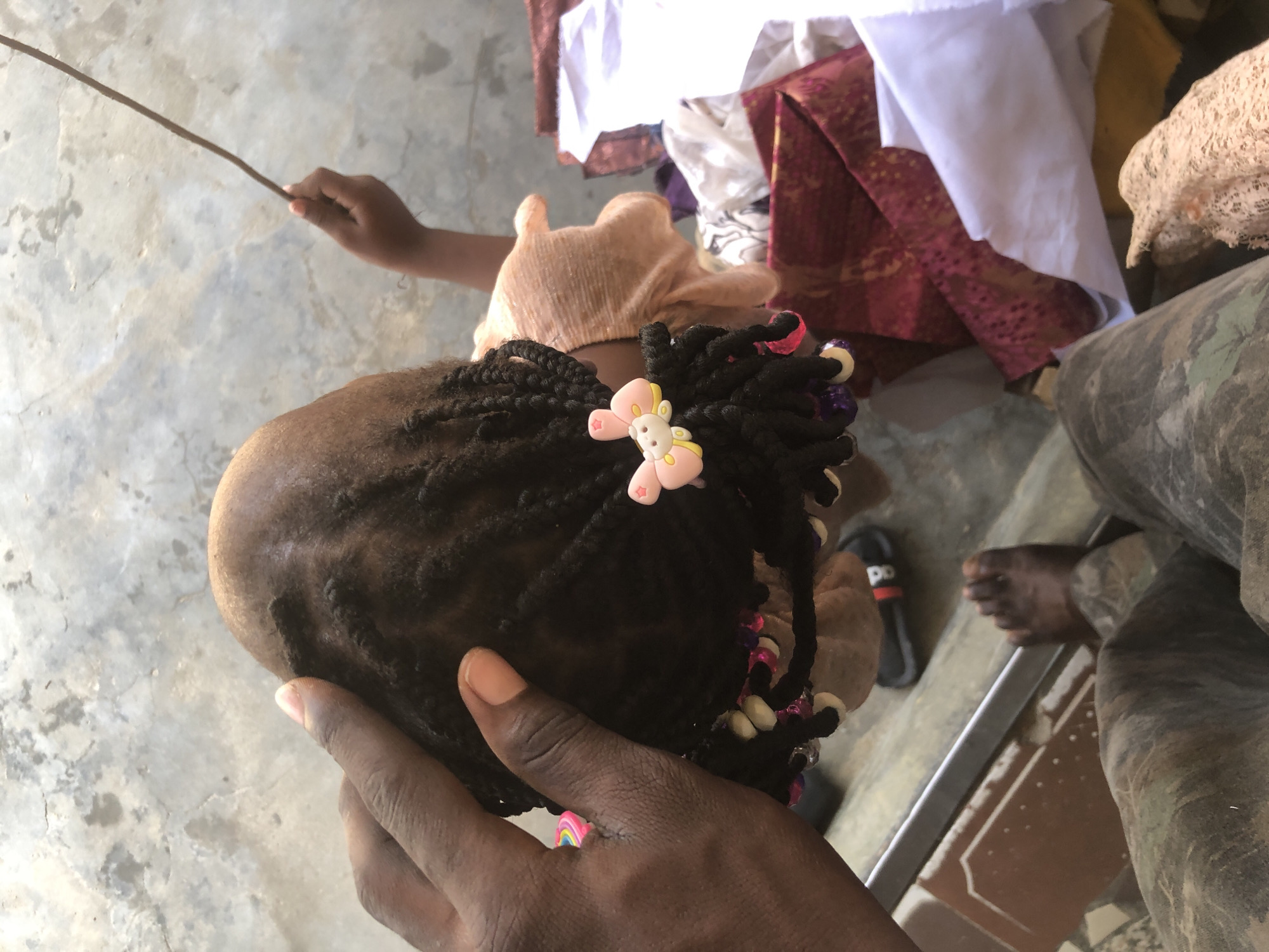 In Ado, Nigeria, this image captures a close-up perspective from above, focusing on a young child's braided hair decorated with colorful beads and a pink bow. A person's dark hand is visible, gently touching the child's scalp. To the right, a pile of fabric, including red and white textiles, is present. The child is wearing a light-colored, ruffled shirt. In the background, a concrete surface with cracks is visible. A thin stick is held by a child's hand extending from the right side of the frame. A person's feet, clad in camouflage trousers, are in the foreground.