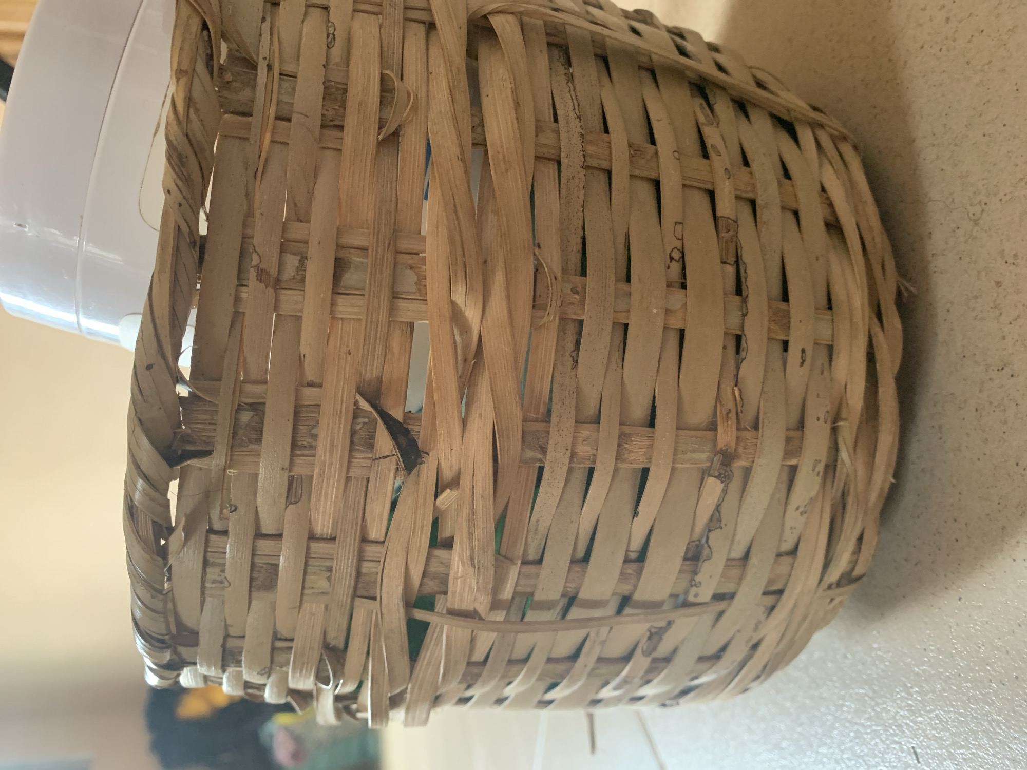 A close-up, eye-level shot shows the side of a round, woven bamboo basket filled with items. The basket is constructed from light brown, unfinished bamboo strips woven in an overlapping pattern. The top rim of the basket is also woven and appears slightly frayed in places. A portion of a white plastic lid from an item inside the basket is visible at the top. The basket sits on a light-colored, tiled floor. In the blurred background, a dark-haired person with a yellow garment is visible. The lighting suggests an indoor setting with natural light illuminating the scene from the side.