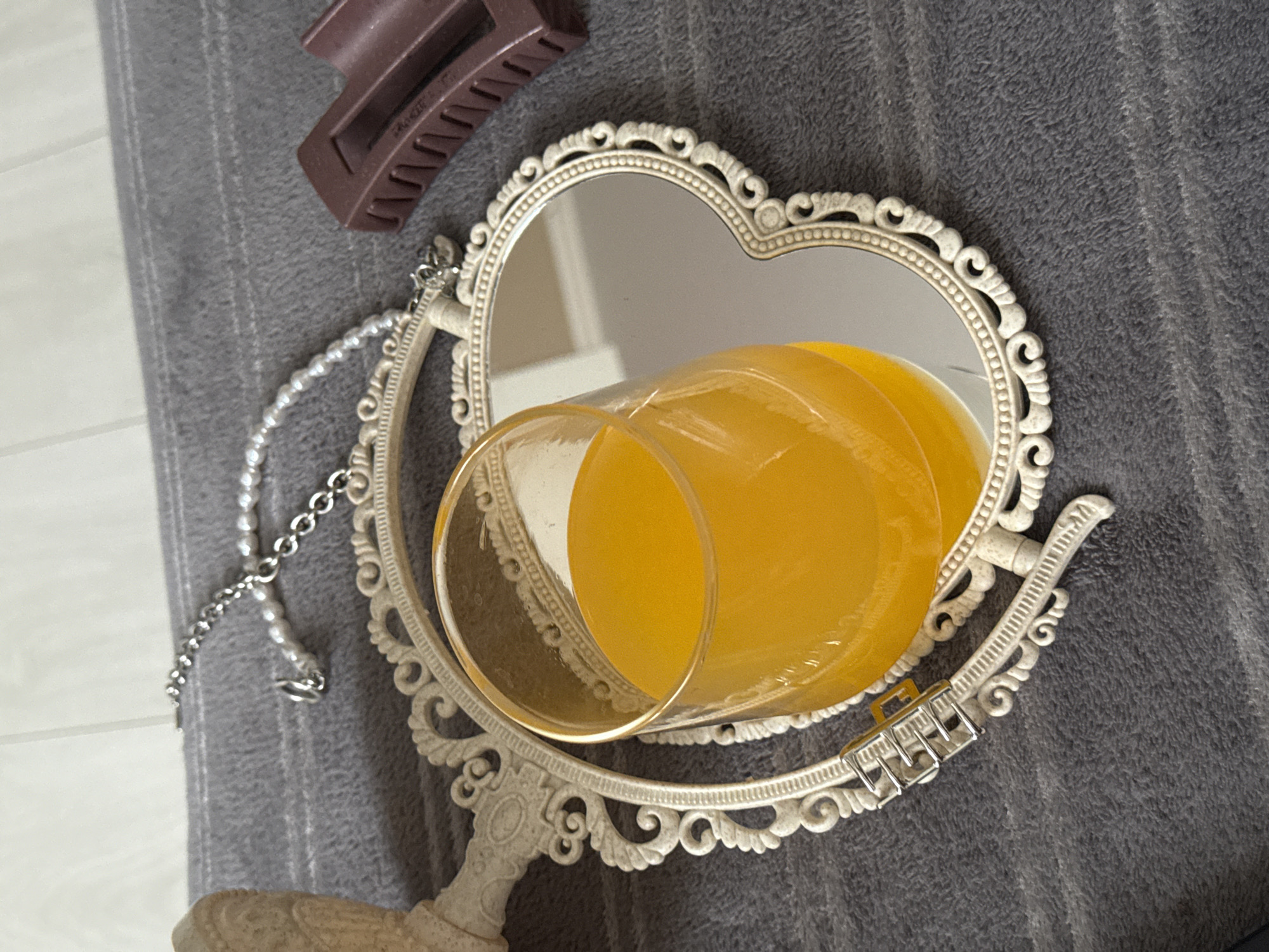 The image shows a still life arrangement. A glass of orange juice is lying on its side within the heart-shaped mirror. A silver necklace is draped near the mirror's left side, and a brown hair clip sits at the top. The scene is set against a grey fabric backdrop. The city and specific location are unknown.