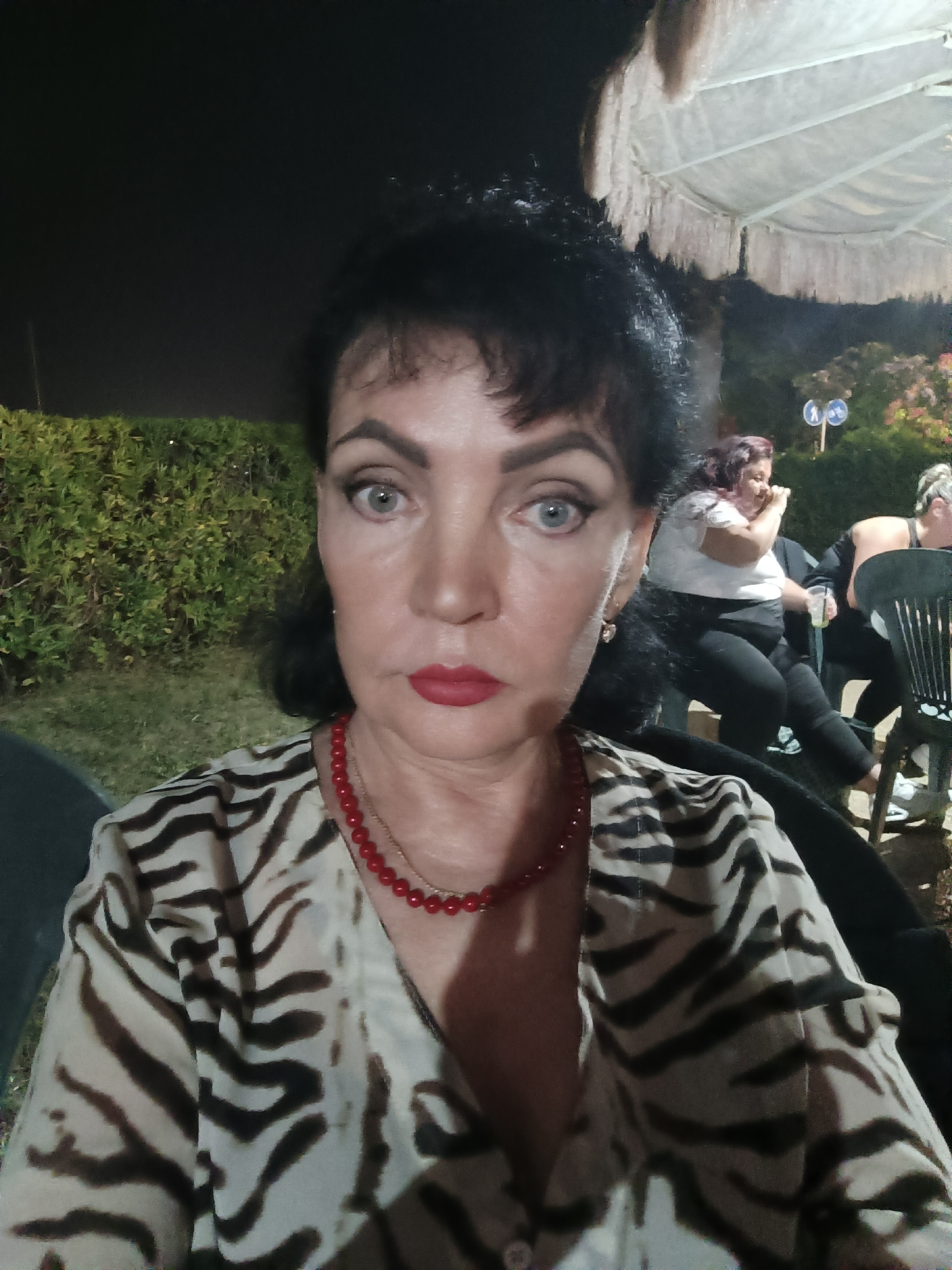 This is a selfie of a woman with dark hair, blue eyes, and red lipstick, wearing a tiger-print shirt and a red beaded necklace. She is outdoors at night, with a hedge and trees in the background. Other people are visible in the background, sitting at tables. The location is identified as Giulianova, Italy.