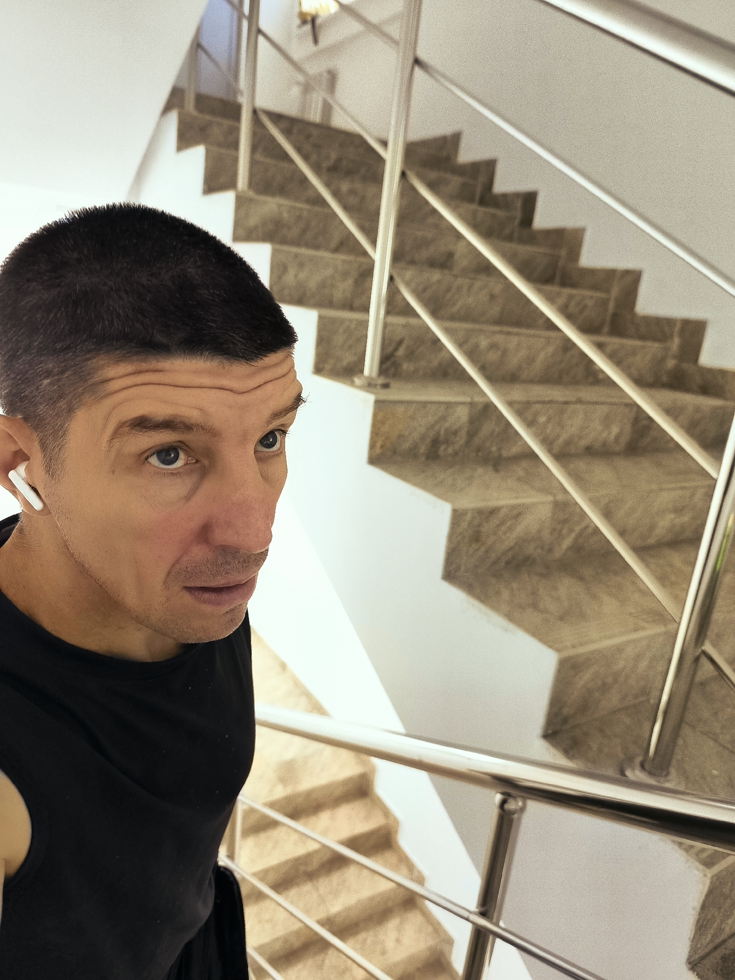A close-up, high-angle shot shows a man wearing a black tank top and white earbuds, looking to his right with a slightly raised eyebrow and his mouth slightly open. He is positioned in the foreground, with a staircase and railing occupying the background. The staircase is made of stone tiles and has a metal railing. The lighting suggests an indoor environment, likely a building with multiple floors. The man's skin appears to be fair, and his hair is short and dark. The overall tone of the image is casual, potentially capturing a moment of pause or exertion. The location in Qarağandı city administration, Kazakhstan is not directly discernible from the visual elements of the image.