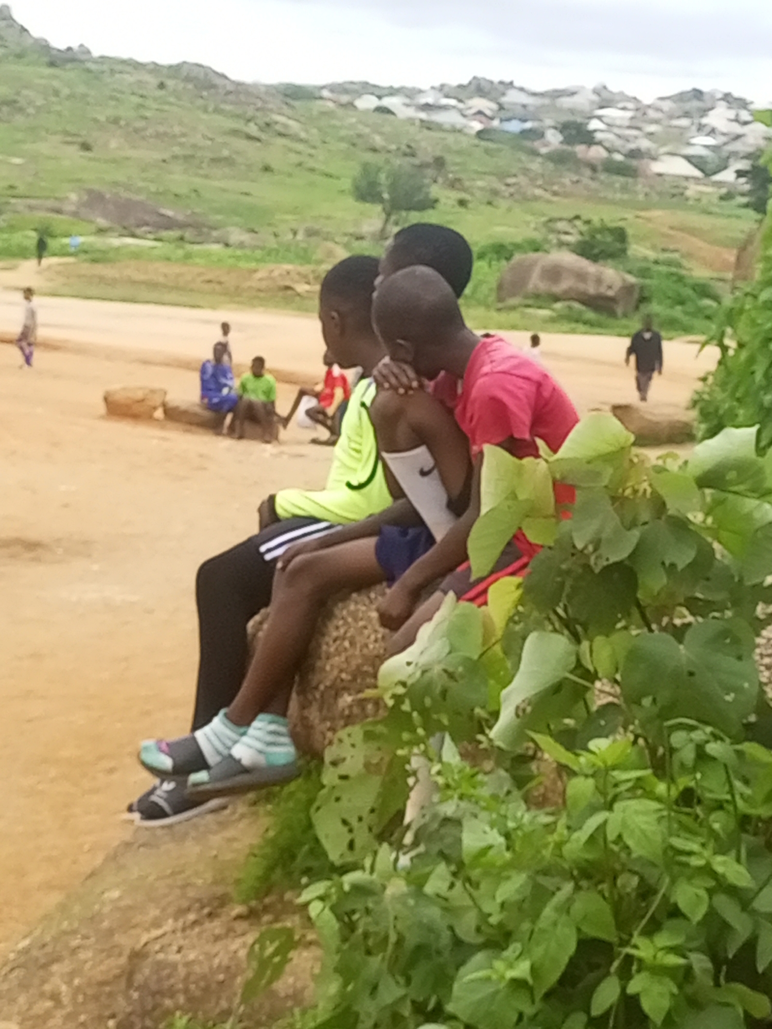 In Jos, Nigeria, three young boys are sitting together on a large rock formation in a rural or semi-rural setting. The boy on the left is wearing a bright yellow long-sleeved shirt and black pants with white stripes, with sandals over striped socks. The middle boy is wearing a white sleeveless shirt and dark shorts, with sandals over socks. The boy on the right is wearing a pink t-shirt and dark shorts. They are sitting close together, with the middle boy appearing to rest his head on the boy to his right. The background shows a dusty, uneven terrain with scattered vegetation and rocks, and a cluster of buildings in the distance, possibly a settlement on a hillside. The sky is overcast.