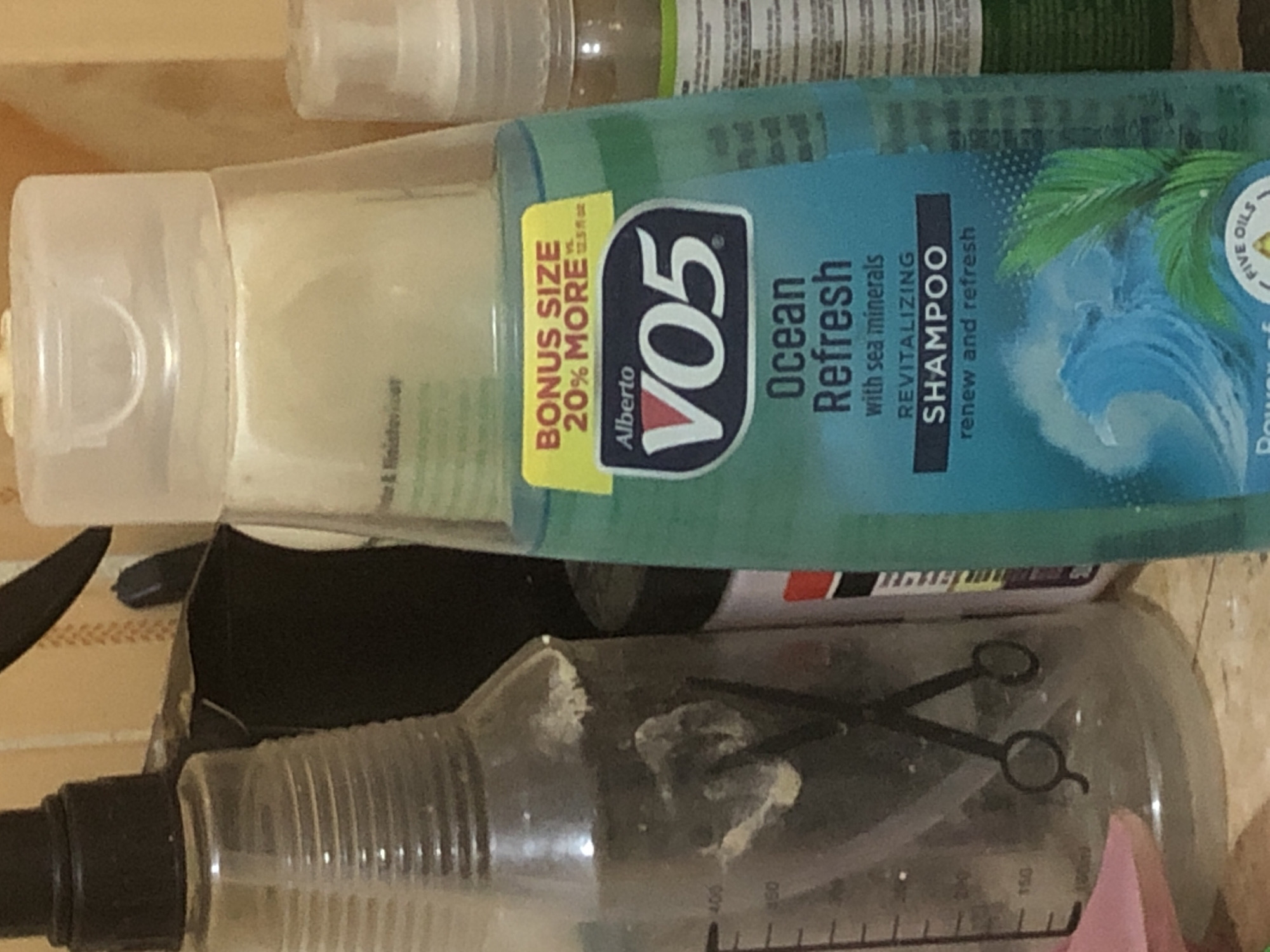 This is a close-up, medium shot of several bottles and containers on a shelf or surface, with a wall in the background. The lighting is somewhat dim and warm, casting shadows.
The most prominent object is a clear plastic bottle of Alberto VO5 Ocean Refresh shampoo, which is partially in focus and takes up the right side of the frame. It has a light blue liquid and features a yellow sticker with "BONUS SIZE 20% MORE" and the VO5 logo. The bottle has a white cap and some text is visible on its label, including "Ocean Refresh," "with sea minerals," and "REVITALIZING SHAMPOO."
To the left of the shampoo bottle is a clear plastic spray bottle with a black spray nozzle. This bottle appears to contain a clear liquid and has markings indicating volume measurements up to 400. Inside this bottle, two black scissors are visible, crossed over each other. There are some whitish residue or contents visible on the inside of this bottle.
Behind these bottles, part of another bottle with a white cap and a translucent body is visible, likely a lotion or conditioner bottle. Further in the background, a wall with a textured pattern, possibly wallpaper or painted tiles, can be seen. The colors of the wall are muted, in shades of beige and brown, with a patterned design.
The image does not contain any information about the city of Kabong, Nigeria, or any specific details about its location within a city. The scene appears to be an indoor setting, likely a bathroom or personal care area, given the nature of the products. There are no people or actions depicted in the image.