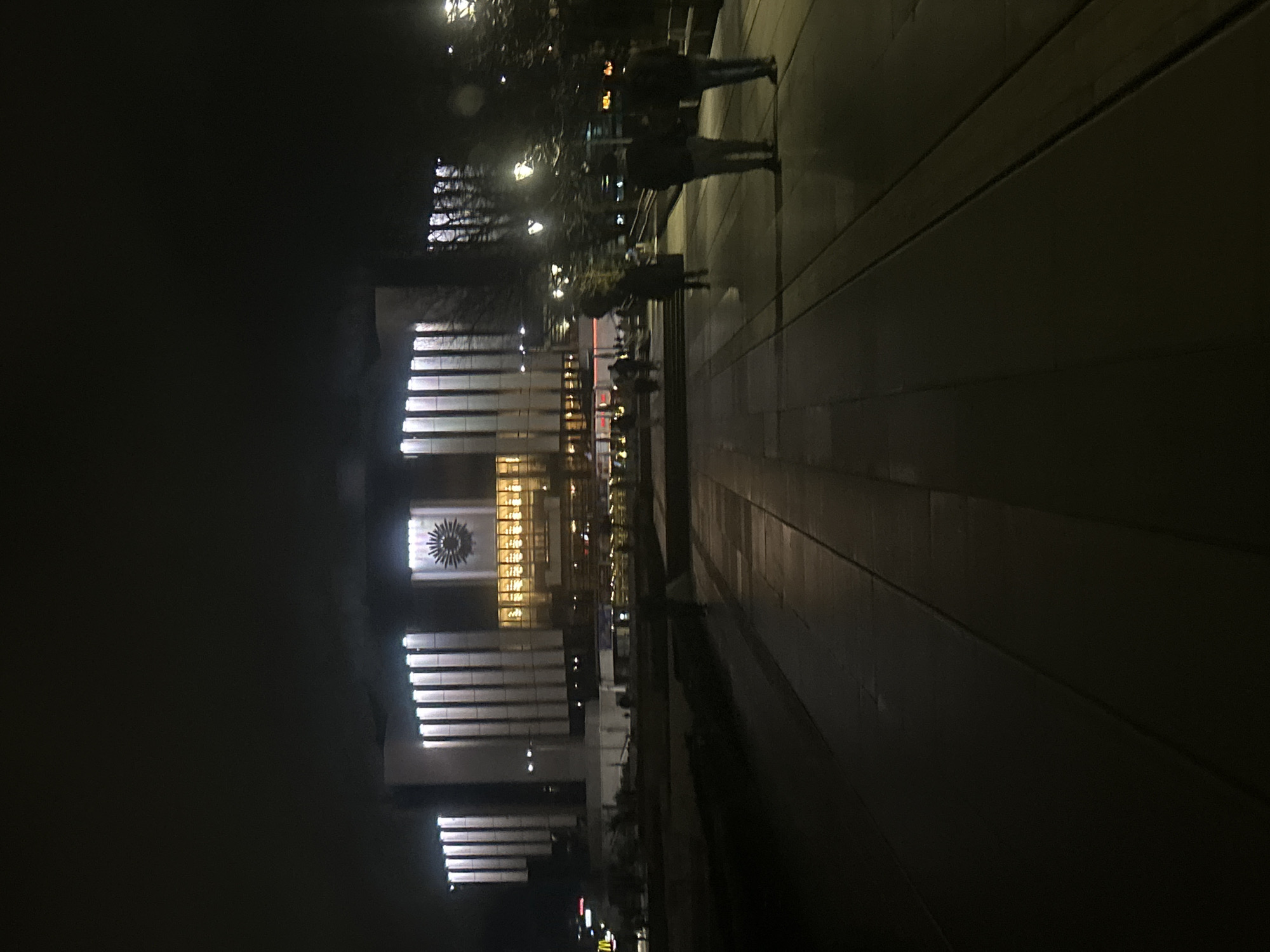The image shows a nighttime view of the National Palace of Culture (NDK) in Sofia, Bulgaria. The building is illuminated, with distinctive vertical lines and a central design element. People are walking on the pavement in the foreground. The environment is dark, with limited ambient lighting.