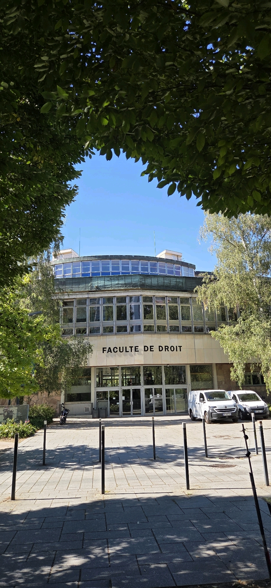 The image shows a large building with the words "FACULTE DE DROIT" inscribed above its entrance. The building is modern, with a circular upper section featuring a band of large windows and a green trim. Below this, the main facade is also covered in windows, framed by stone and metal. The entrance consists of large glass doors. In front of the building, there is a paved walkway with several dark bollards. Two white vans are parked to the right of the entrance. To the left of the entrance, a motorcycle is parked near some bushes. The scene is framed by lush green foliage from trees in the foreground, with a clear blue sky visible through the leaves and above the building. The lighting suggests it is a sunny day, casting shadows on the pavement.