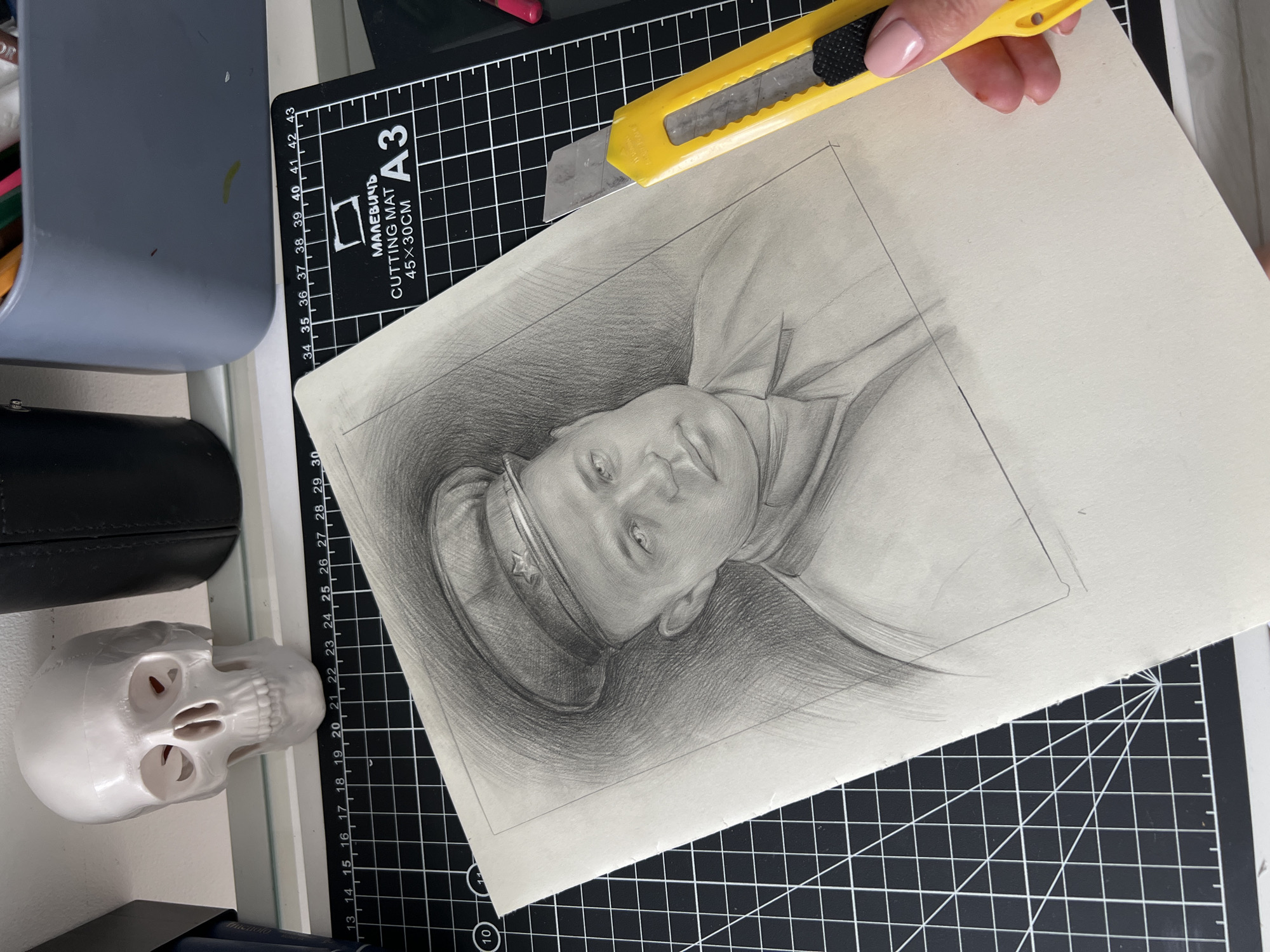 The image displays a close-up, overhead shot of a detailed pencil portrait of a man in uniform, resting on a black cutting mat with grid lines and measurements. The portrait is positioned in the lower center of the frame, with a portion of the man's face and shoulder visible. The man is wearing a cap with a star emblem, and his expression is serious. To the right of the portrait, a yellow utility knife is held by a person with manicured pink nails, its blade poised near the edge of the paper. In the background, a white skull figurine is visible on the left, and a gray plastic container filled with art supplies is on the right, suggesting an art studio or workspace. The overall scene conveys an artist's working environment.