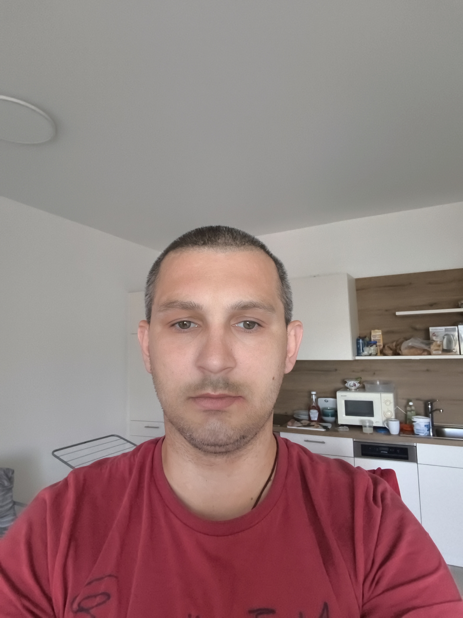 A medium shot shows a man with short brown hair and light skin, wearing a red t-shirt, looking directly at the camera. He has a light stubble and his expression is neutral. The background suggests an indoor setting, possibly a kitchen. There are white cabinets and a wooden shelf with various items, including a microwave and kitchenware. A sink and countertop are also visible. The room appears to be well-lit, with a ceiling light fixture and natural light entering from an unseen source. The image does not contain any specific landmarks or elements that would definitively place it in Wolgast, Germany.