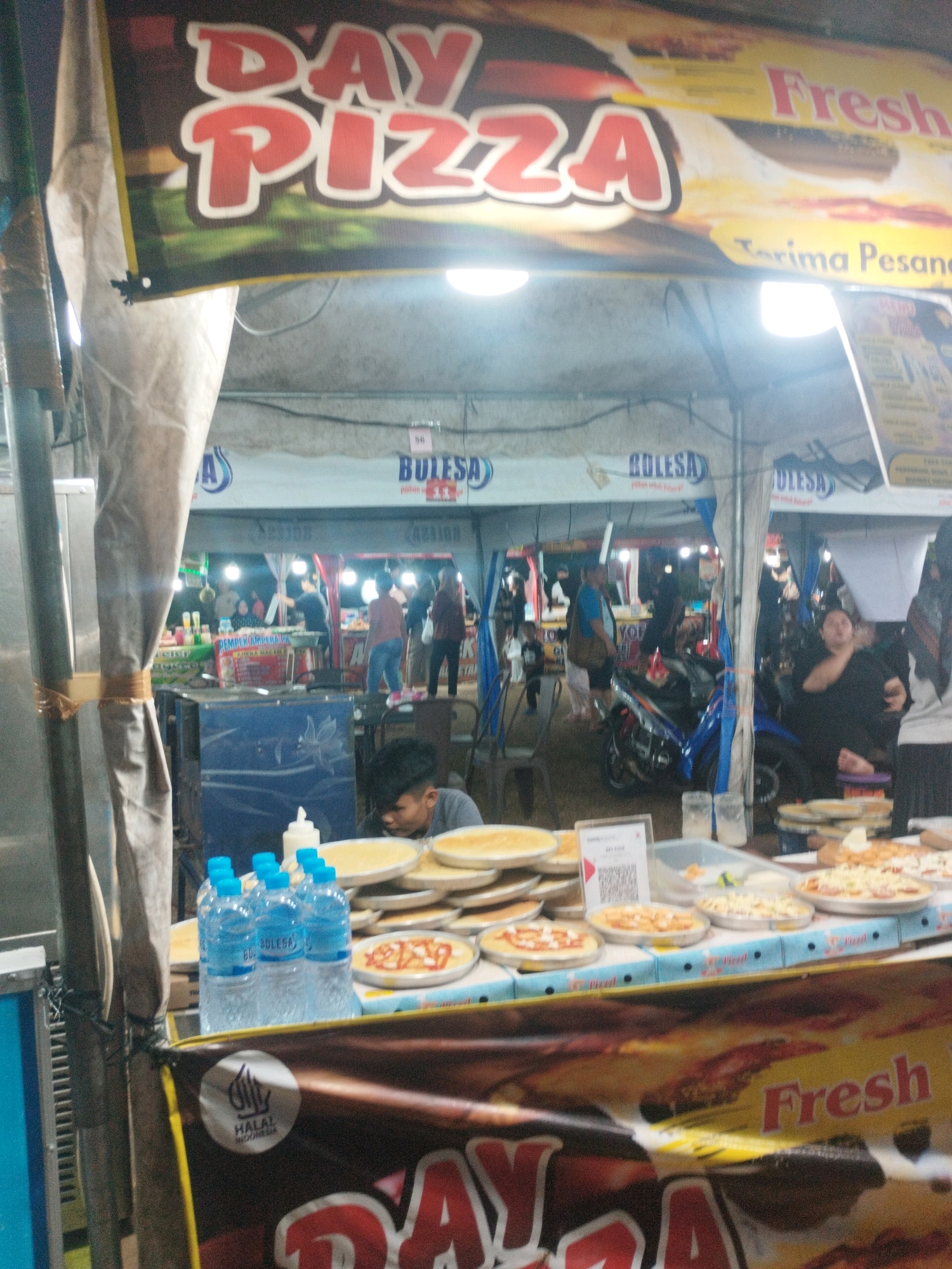 The image depicts a food stall, possibly a pizza stand, in what appears to be an outdoor market or street food setting in Parit Padang, Indonesia. A prominent banner displays "DAY PIZZA" in large, red, stylized lettering, with "Fresh" also visible in red text. The stall is set up with a table displaying several pizzas, some in baking pans and others already prepared. There are bottles of water and condiments on the table.
In the background, other stalls and people are visible, suggesting a bustling commercial area. One stall in the background has signage that reads "BULESA". There are also glimpses of motorcycles and various individuals, including a young man seated in the foreground, likely associated with the food stall. The overall lighting suggests it might be evening or nighttime, with artificial lights illuminating the scene. The presence of a "HALAL INDONESIA" logo on a banner indicates compliance with Islamic dietary laws.