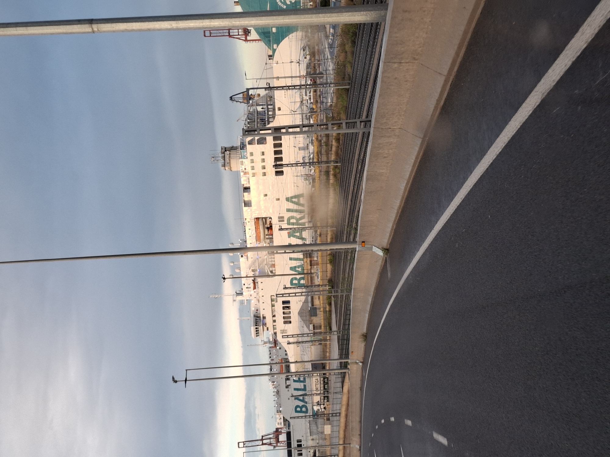 This image depicts a view in the port area of Valencia, Spain. The scene is dominated by the presence of several large ferries, specifically those operated by the Baleària company, with the word "BALEÀRIA" prominently displayed on the ships. The ferries are white, and their decks and superstructures are visible. The lighting suggests it is either early morning or late afternoon.
Parallel to the ferries are railway tracks. A road is visible on the right side, with a white line marking the edge of the lane. There are light posts and power lines lining the street.
The sky is pale and appears cloudy. There are glimpses of crane structures and other port infrastructure in the background.