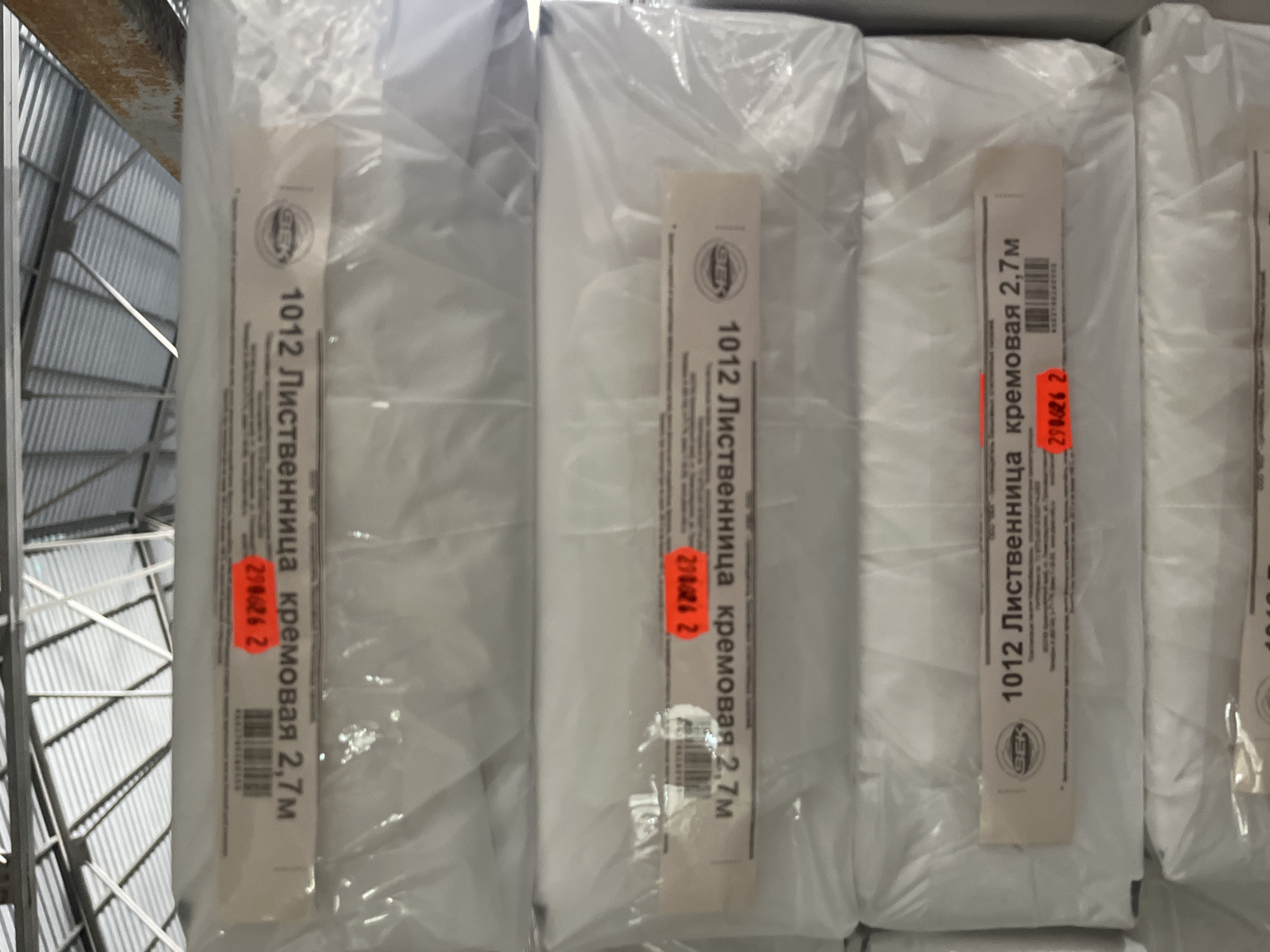 The image shows a close-up, eye-level view of several rolls of material, likely fabric or paper, stored vertically. The rolls are individually wrapped in clear plastic and are arranged in a row, suggesting they are part of a larger inventory. Each roll has a label attached to its front, providing product information. The labels are predominantly white with black text.
The visible text on the labels indicates the product is "1012 Лиственница кремовая 2,7м," which translates to "1012 Larch cream 2.7m." This suggests the product is a cream-colored material, possibly a textile or a decorative paper, with a width of 2.7 meters and a model number of 1012. Some labels also feature a circular logo with the letters "GEK" or "SEK" inside. Additionally, some rolls have red price tags or labels with a numerical code, such as "290626 2" and "294026 2."
The background of the image reveals a warehouse or storage environment. Metal shelving units with a ribbed metal surface are visible on the left side, suggesting a systematic storage solution. The lighting appears to be artificial, typical of an indoor commercial space, and there are no indications of natural light or weather conditions. The overall impression is one of organized retail or wholesale stock. The image is taken from a straight-on perspective, focusing on the product labels and the texture of the plastic wrapping.
