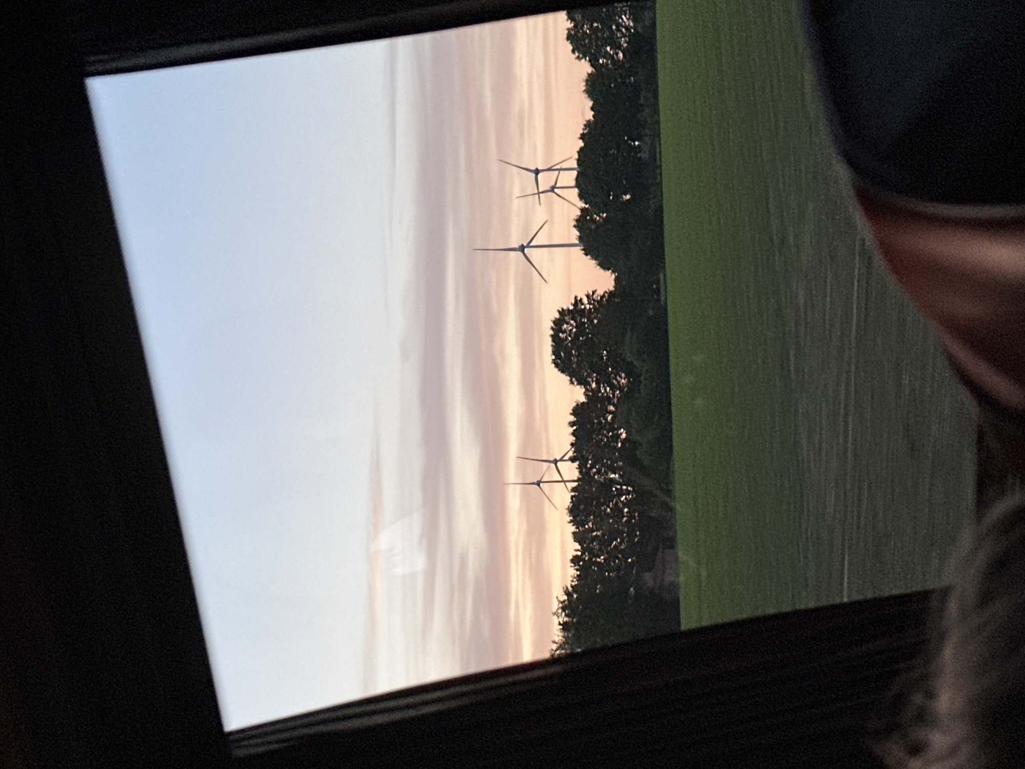 The image, likely taken from inside a vehicle through a window, shows a view of a field with wind turbines at dusk or dawn. The sky is a gradient of light blue to pale orange. There are at least three wind turbines visible in the distance, partially obscured by trees. The field is green. The location is near Harsefeld, Germany, indicated by the presence of wind turbines, a common feature of the landscape in that area. The vehicle's interior is dark and out of focus. A person's head is visible in the lower right corner of the image.