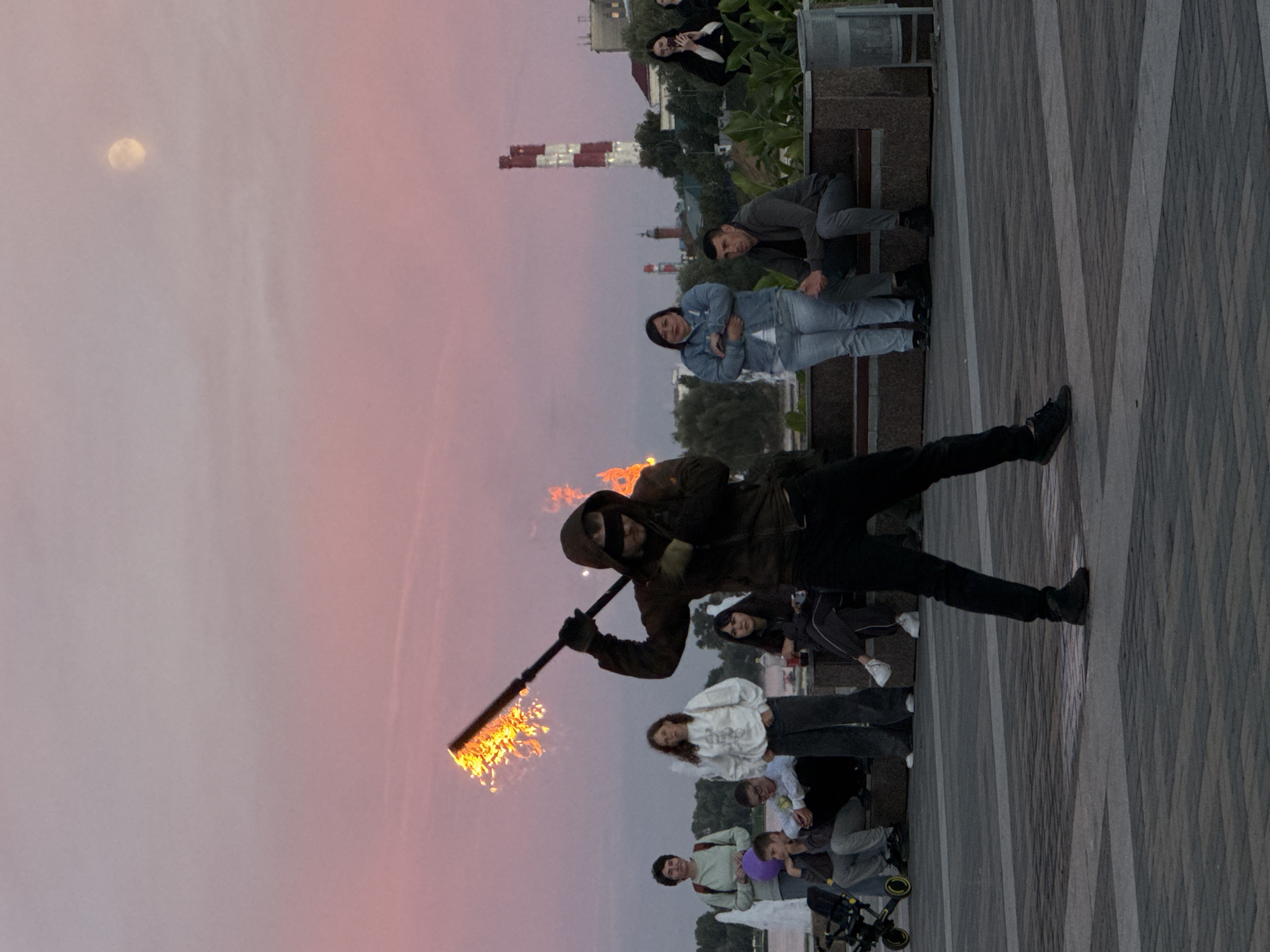 A group of people are gathered outdoors as a performer juggles with a flaming staff. The sky is a soft pink and purple, suggesting either sunrise or sunset, and a full moon is visible high in the sky. The performer, wearing a dark hooded jacket and gloves, stands in the foreground with the flaming staff held horizontally, its flames licking upwards. A small crowd of onlookers, some standing and some seated on a bench, watch the performance. Among the spectators are individuals of various ages, some dressed casually in jeans and hoodies. In the background, there are industrial buildings with red and white striped chimneys. The ground is paved with a textured, grey material. The overall scene has a theatrical and energetic feel, with the dramatic fire performance set against a serene sky.