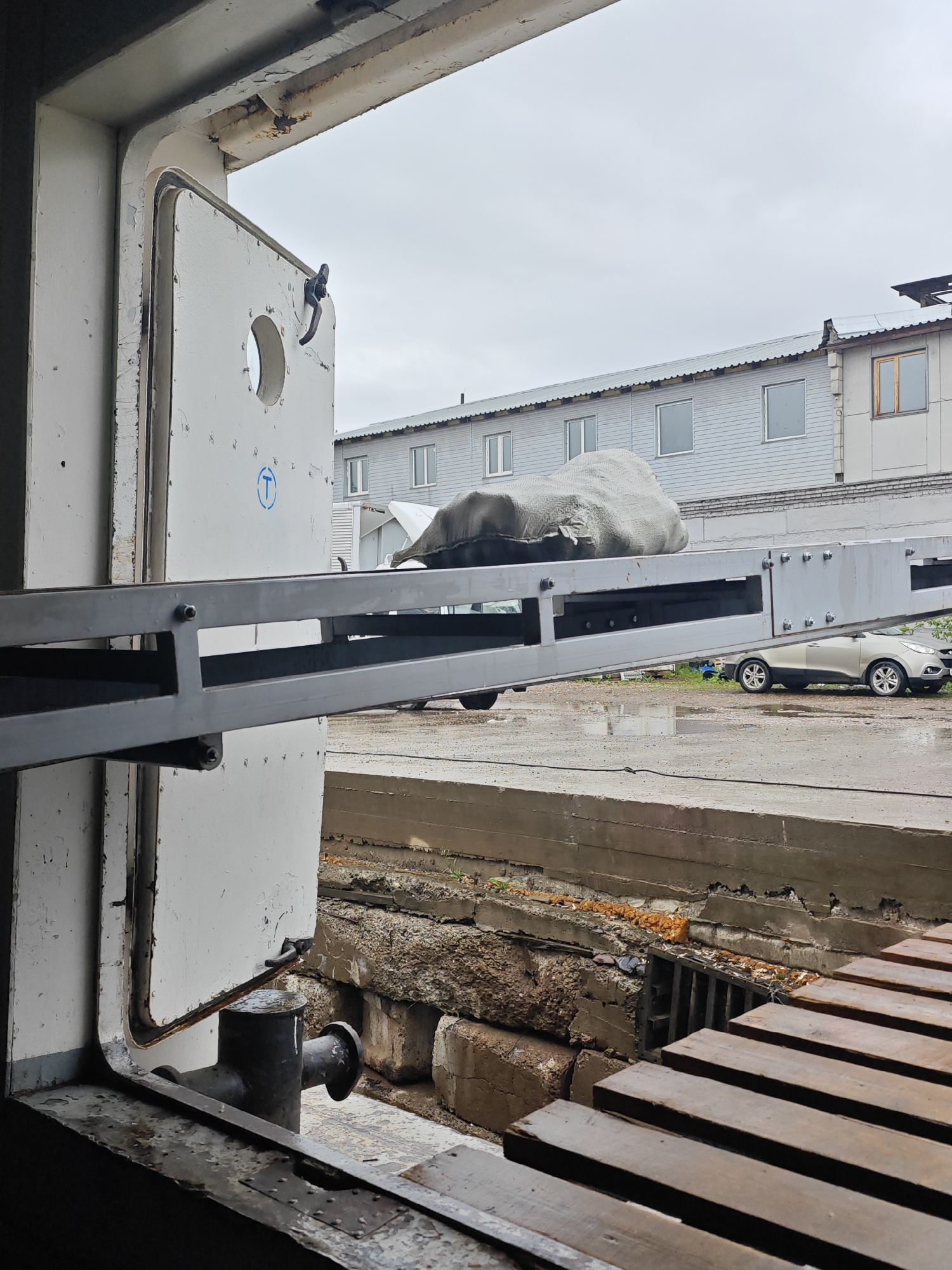 The image depicts the open cargo bay of a vessel, likely a ferry or barge, viewed from the interior. A ramp extends outwards from the vessel, leading to a paved area. A light-colored car is parked on the paved area, partially under the ramp. The car is wet, suggesting recent rain. A large object covered in grey tarp is visible on the ramp. In the background, a long, low building with multiple windows is situated. The sky is overcast and gray. The scene appears to be a port or industrial area. Based on the visual context of a port and industrial buildings, it is possible this location is in Krasnoyarsk, Russia, which has a significant river port on the Yenisei River.
