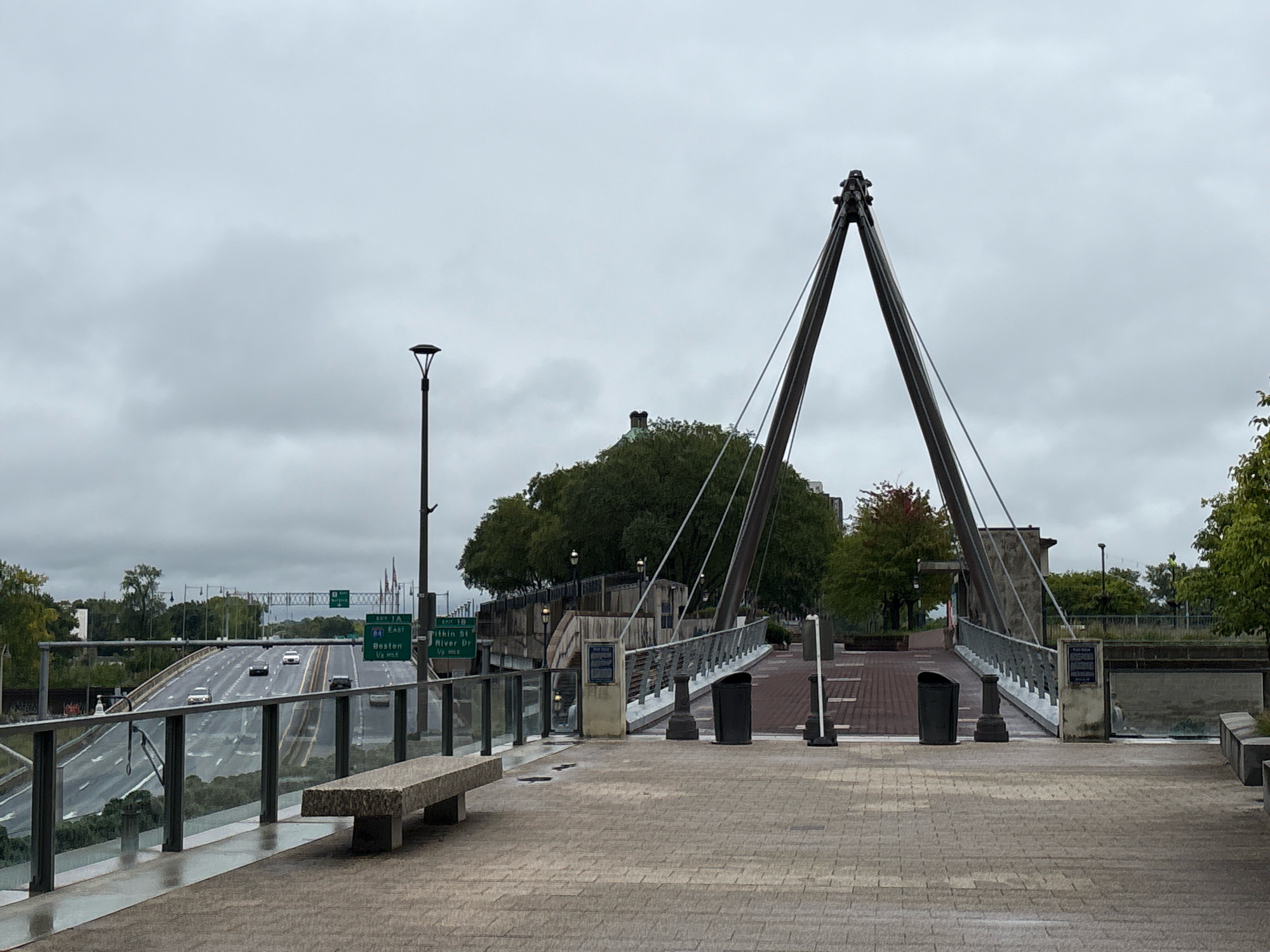 The image depicts an overcast day in Hartford, Connecticut, USA, focusing on a pedestrian bridge that spans over a highway. In the foreground, a paved walkway with a stone bench to the left leads towards the bridge. The walkway is constructed of brick-like pavers. The pedestrian bridge itself features a modern design with a cable-stayed structure, characterized by two angled towers that support the walkway. Several streetlights are visible along the bridge and the surrounding elevated walkway.
To the left of the pedestrian bridge, Interstate 84 is visible, with multiple lanes of traffic and cars driving on it. Green highway signs indicating "EXIT 1A EAST", "84 EAST", "Boston", and "EXIT 1B" are prominently displayed above the highway. The environment suggests an urban setting with a mix of infrastructure and greenery, including trees that line the highway and behind the pedestrian bridge. The sky is uniformly gray, indicating cloudy weather. There are no people visible in the frame, but there are trash receptacles on the elevated walkway of the bridge. The overall scene is one of urban infrastructure and transit, observed from a public walkway.