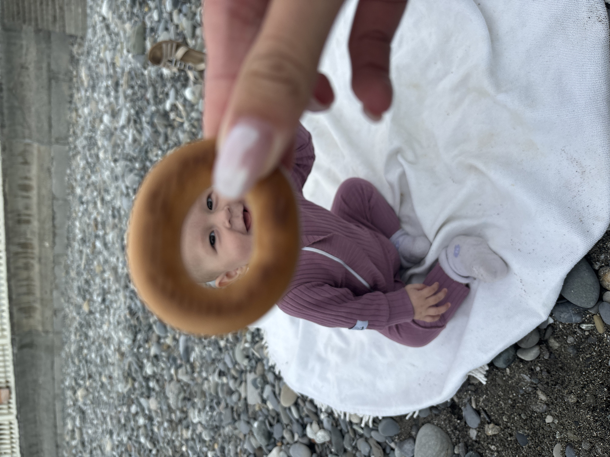 The image shows an infant sitting on a white blanket on a pebble beach. A hand holds a round pastry in front of the baby's face, creating a frame. The baby wears a mauve-colored outfit and white socks. In the background, there's a concrete structure and a pair of sandals. The scene is likely located on a beach in Sochi, Russia, based on the typical pebble beaches and coastal structures found in that area.